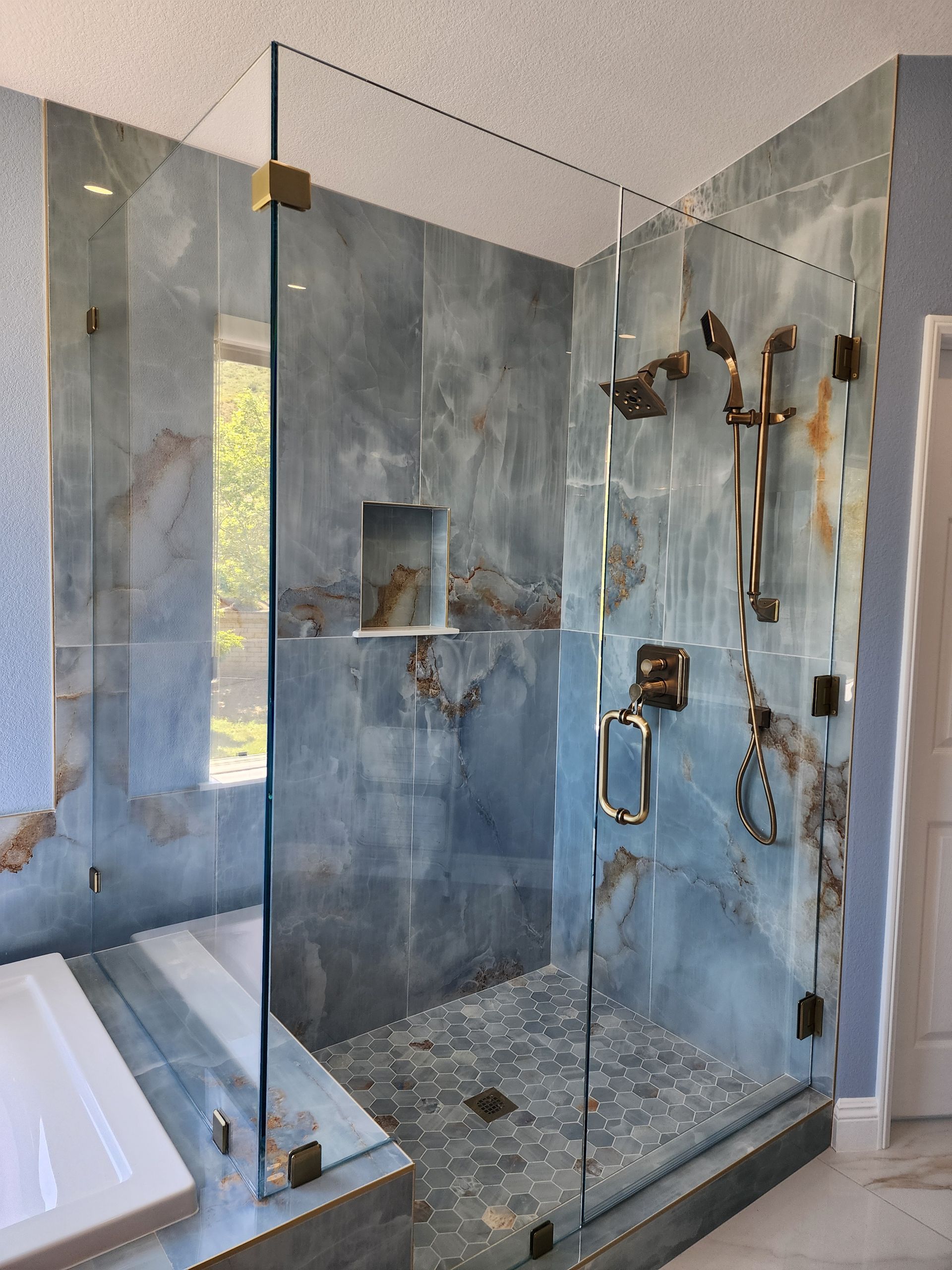 A modern walk-in shower with large blue-gray marble tiles, pebble flooring, and brass fixtures behind a glass door.
