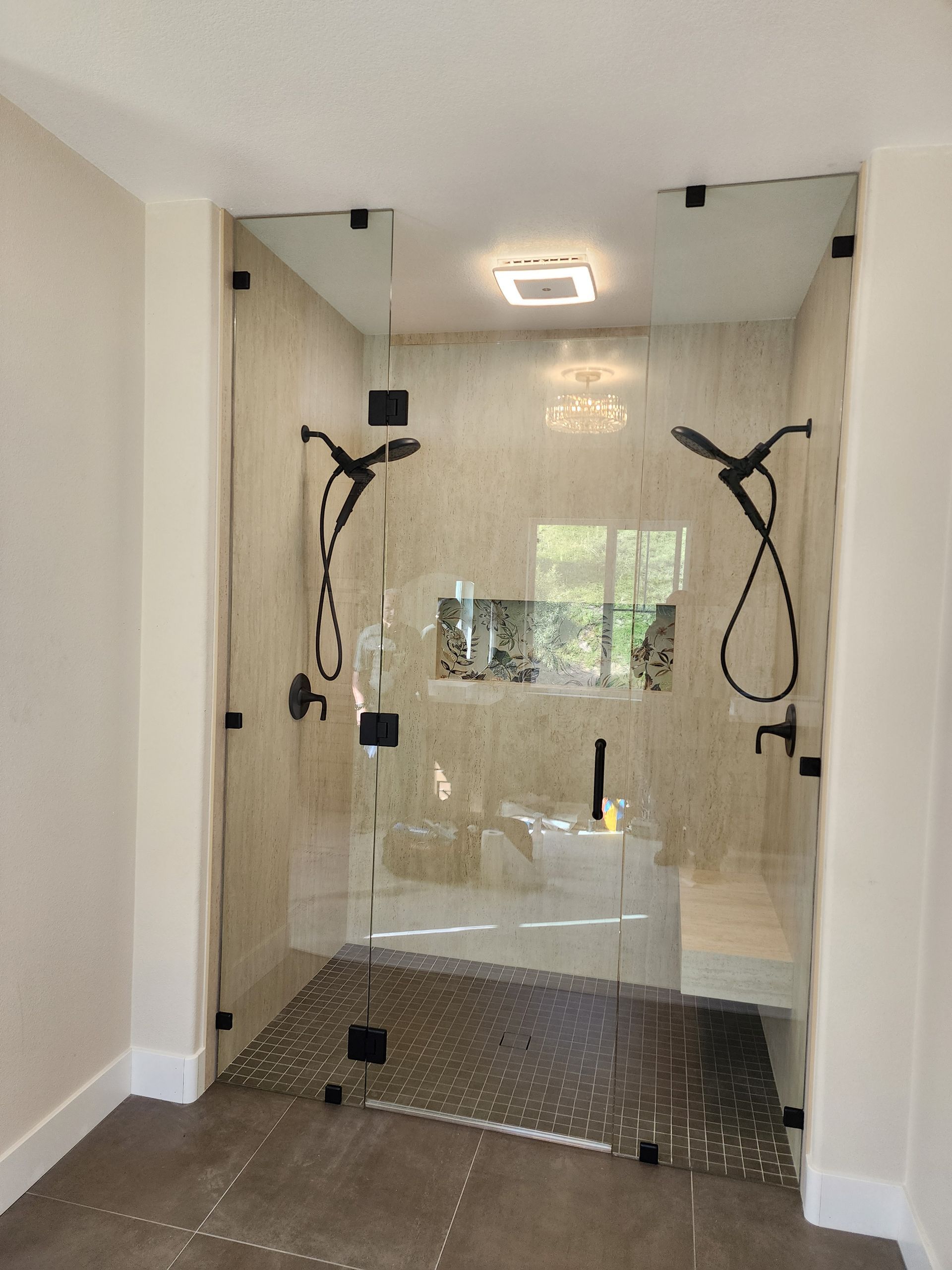 A modern walk-in shower with beige tile walls, glass doors, black hardware, two handheld showerheads, and a built-in bench.