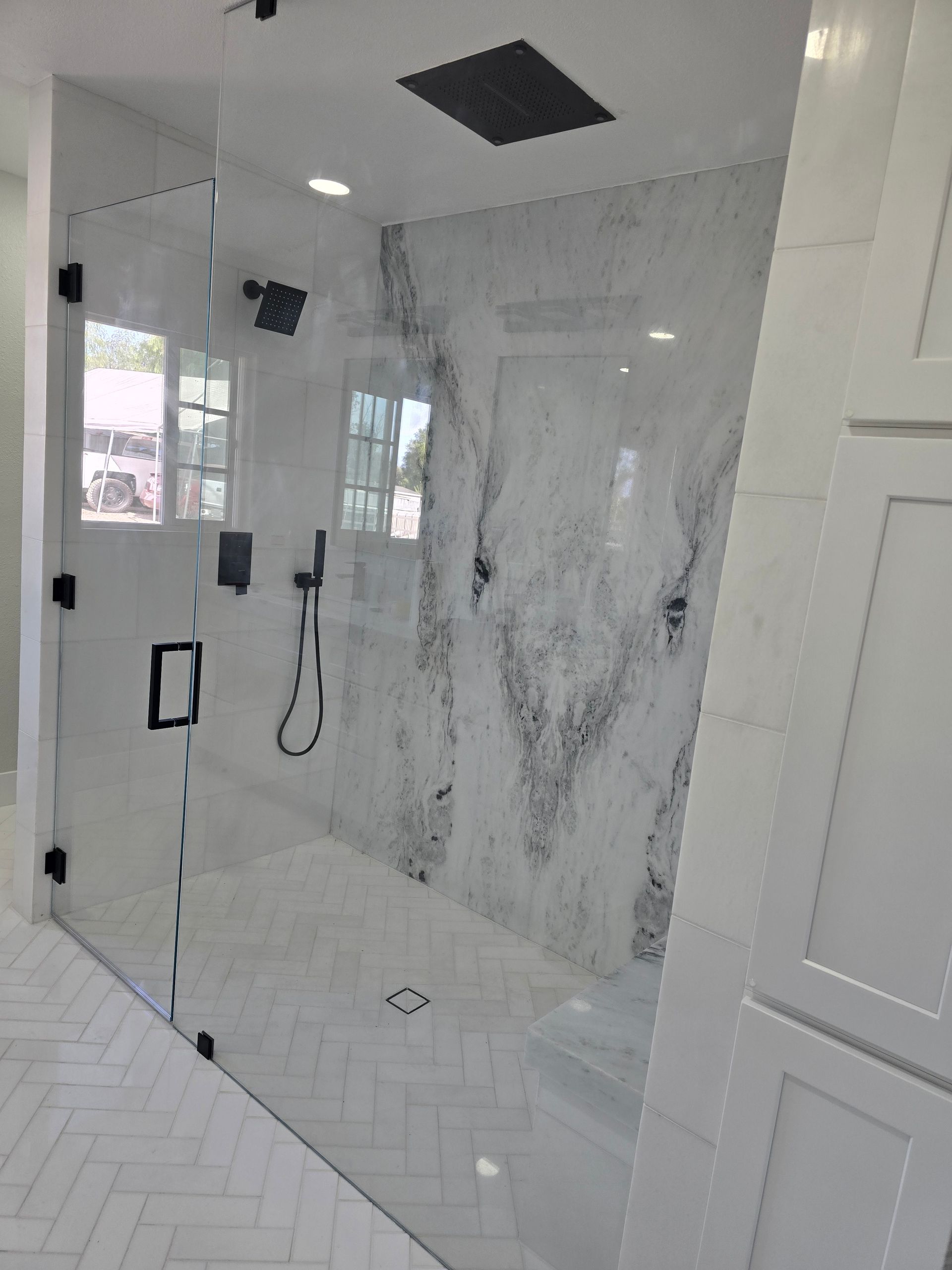 A modern walk-in shower with a large white marble wall, glass door, black fixtures, and a white herringbone tiled floor.