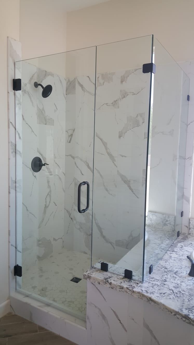 A glass walk-in shower with black hardware, marble-patterned tile walls, and a granite-topped vanity.