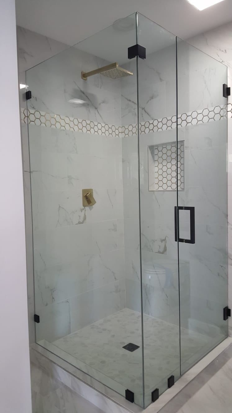 A glass-enclosed walk-in shower with white marble walls, gold hardware, black fixtures, and a decorative tile accent.