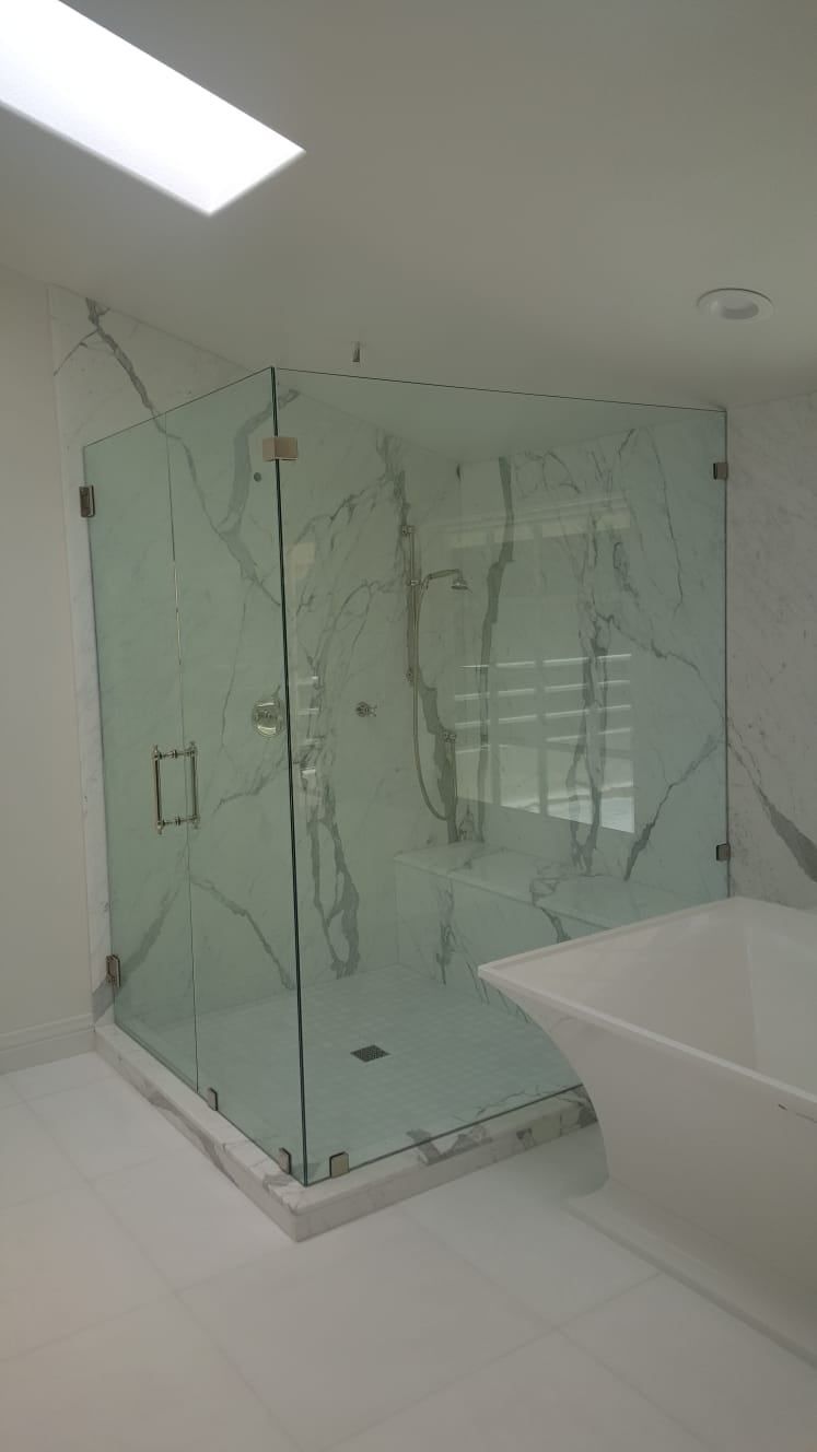 A frameless glass shower enclosure with marble-patterned tile walls, located next to a white freestanding bathtub.