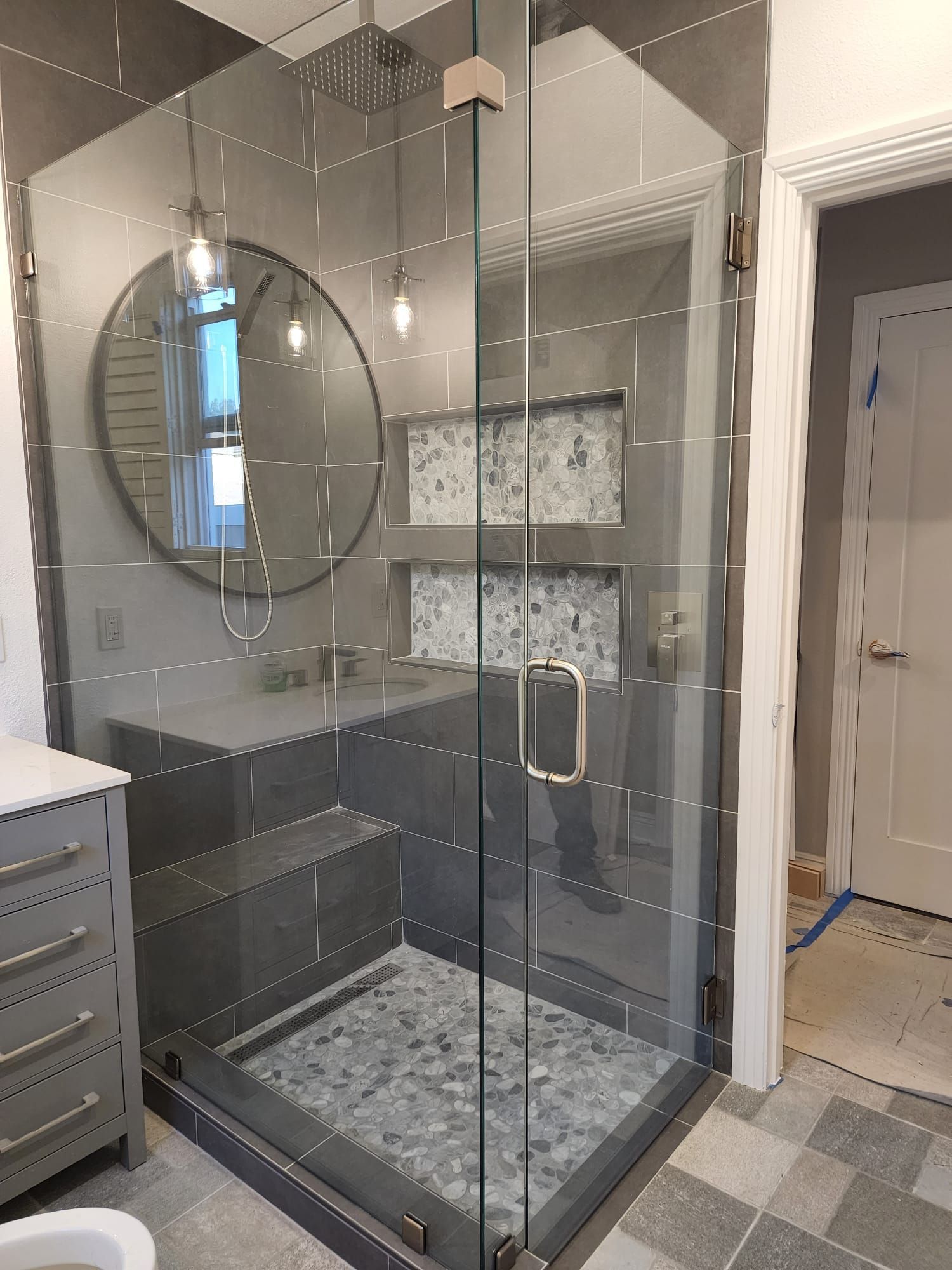 A modern glass shower enclosure with dark grey tiles, a pebble floor, a built-in bench, and dual recessed wall shelves.