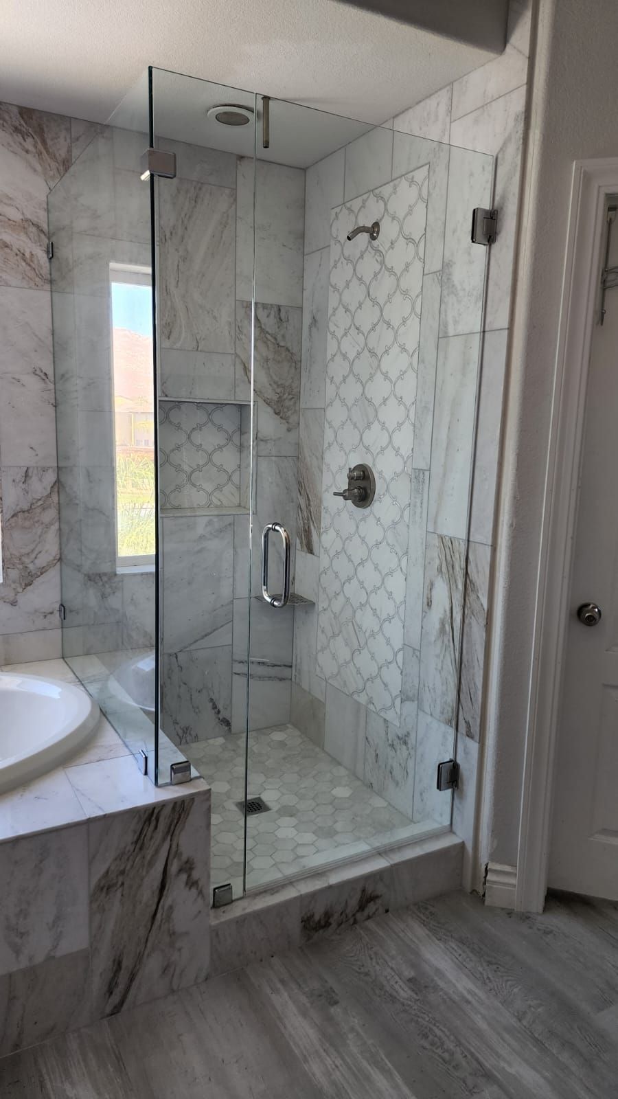 A walk-in glass shower with marble-style tile walls, a patterned accent wall, and a silver shower head.