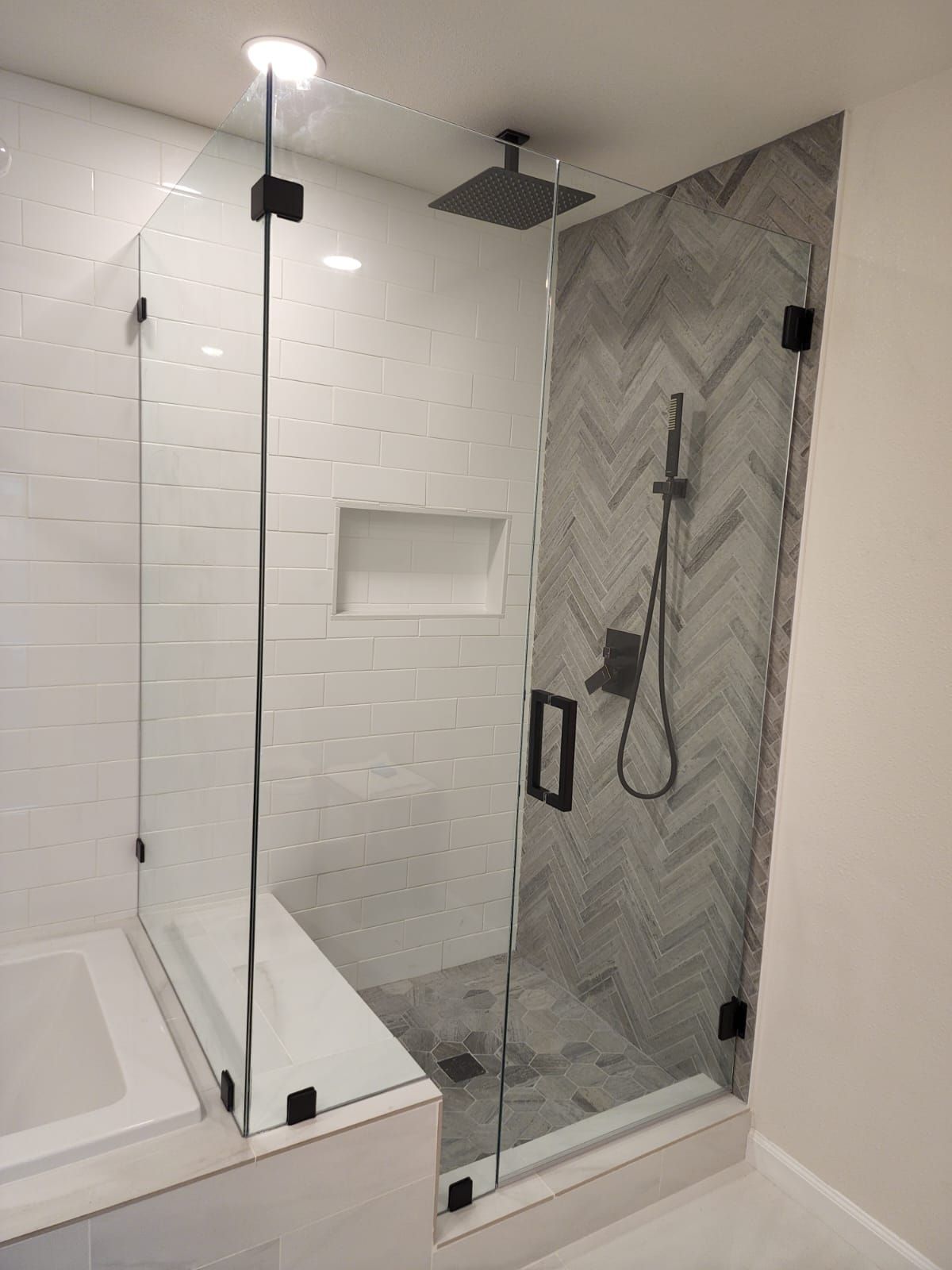 A modern walk-in shower with a glass door, black hardware, white subway tile walls, and a gray herringbone accent wall.