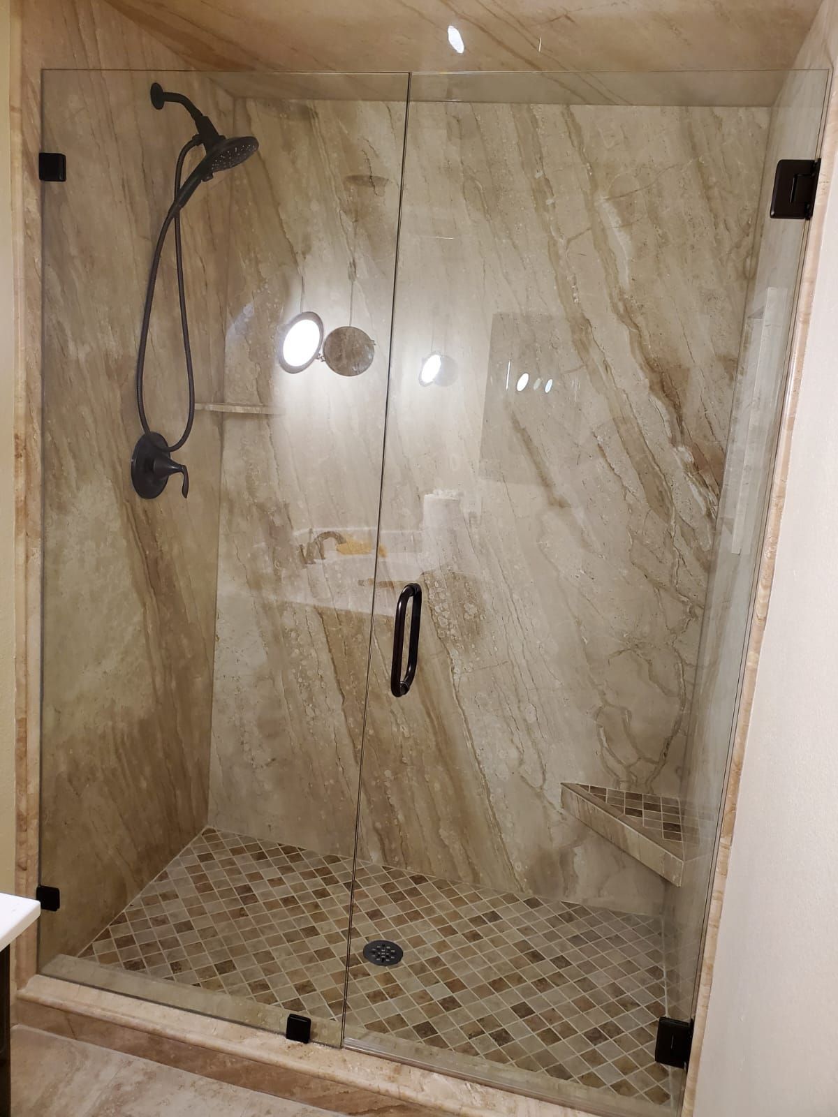A walk-in shower with beige marble-look tiles, a patterned tile floor, a glass door, and dark metal hardware.