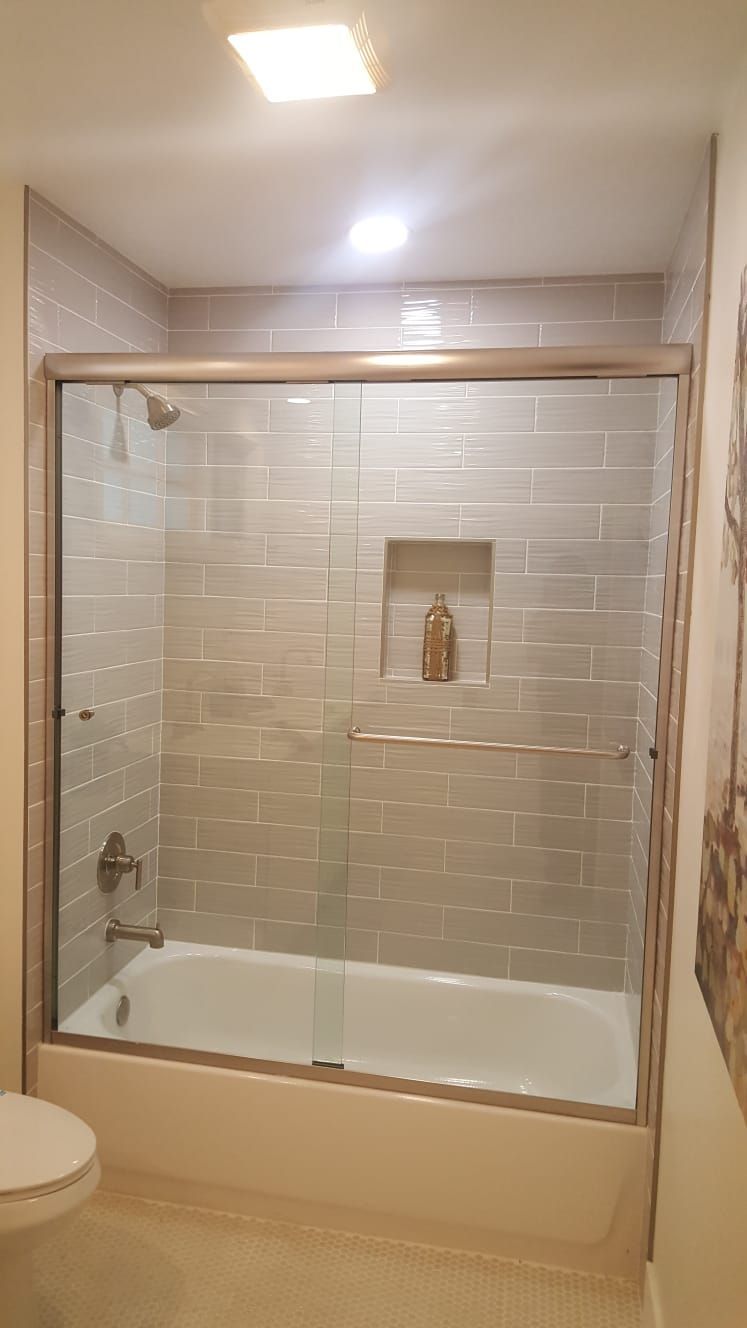 A modern bathroom with a white bathtub, glass sliding shower doors, and light gray subway tile walls with a built-in niche.