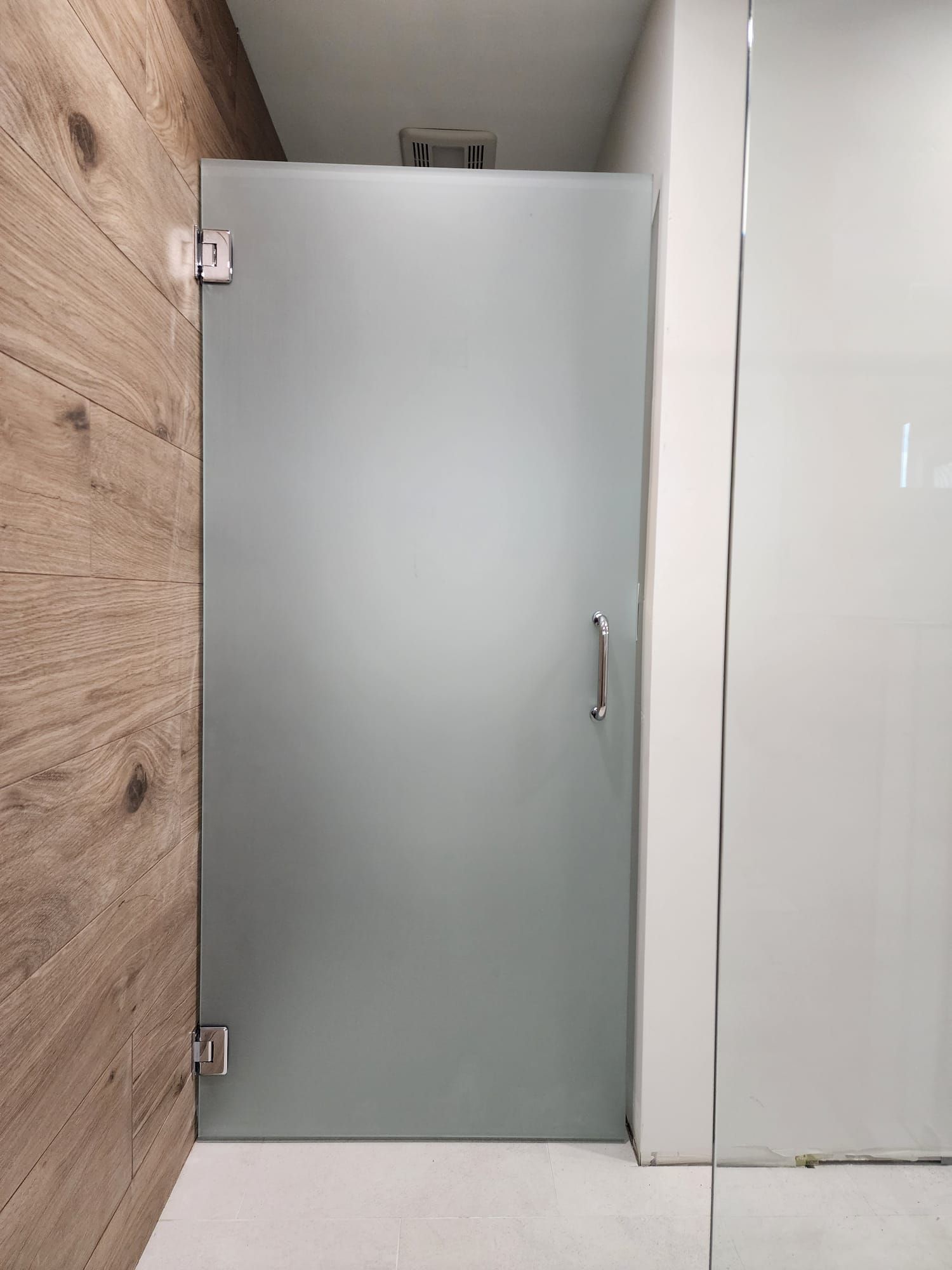 A frosted glass shower door with silver hinges and handle, positioned between a wood-paneled wall and a white wall.
