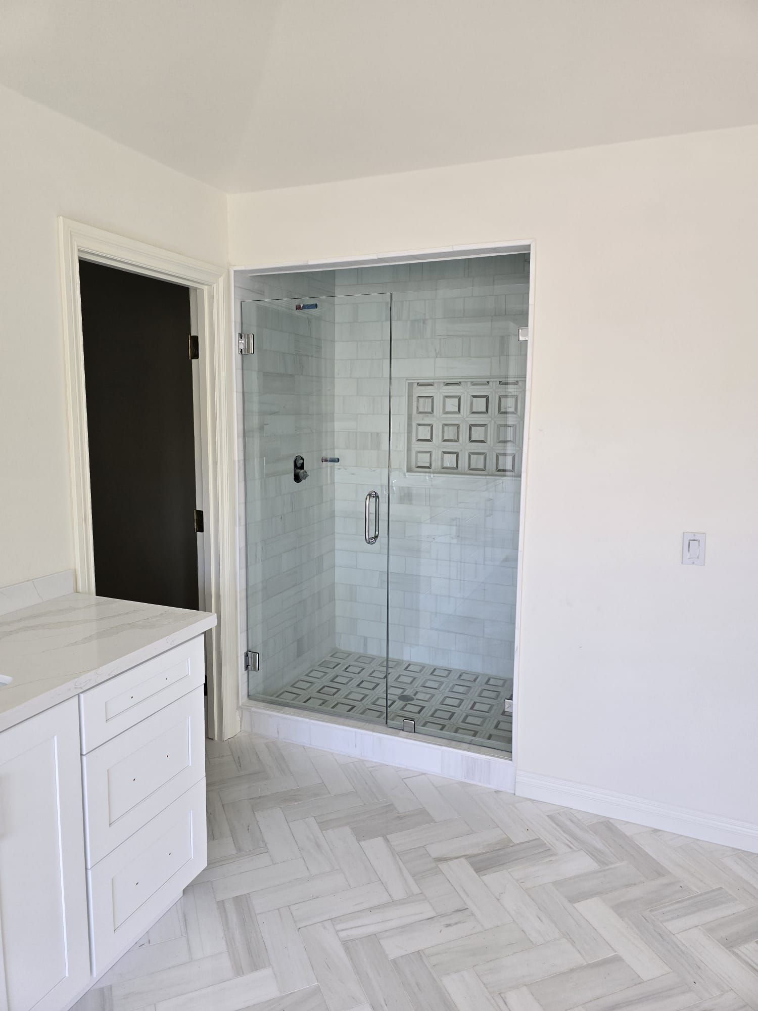 A modern bathroom featuring a white vanity, herringbone floor tiles, and a walk-in glass shower with marble-style tiles.