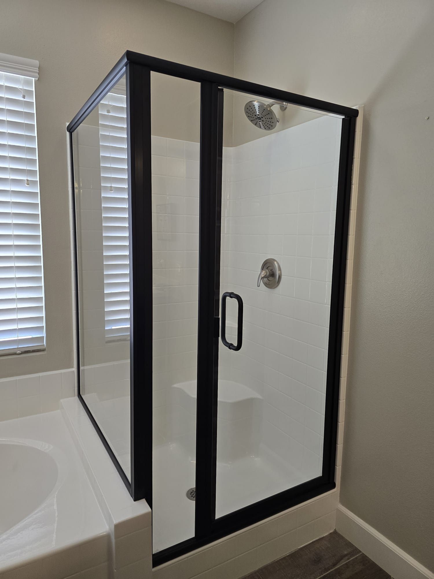 A corner shower stall with a black frame and glass doors, situated in a bathroom with a nearby bathtub and window blinds.