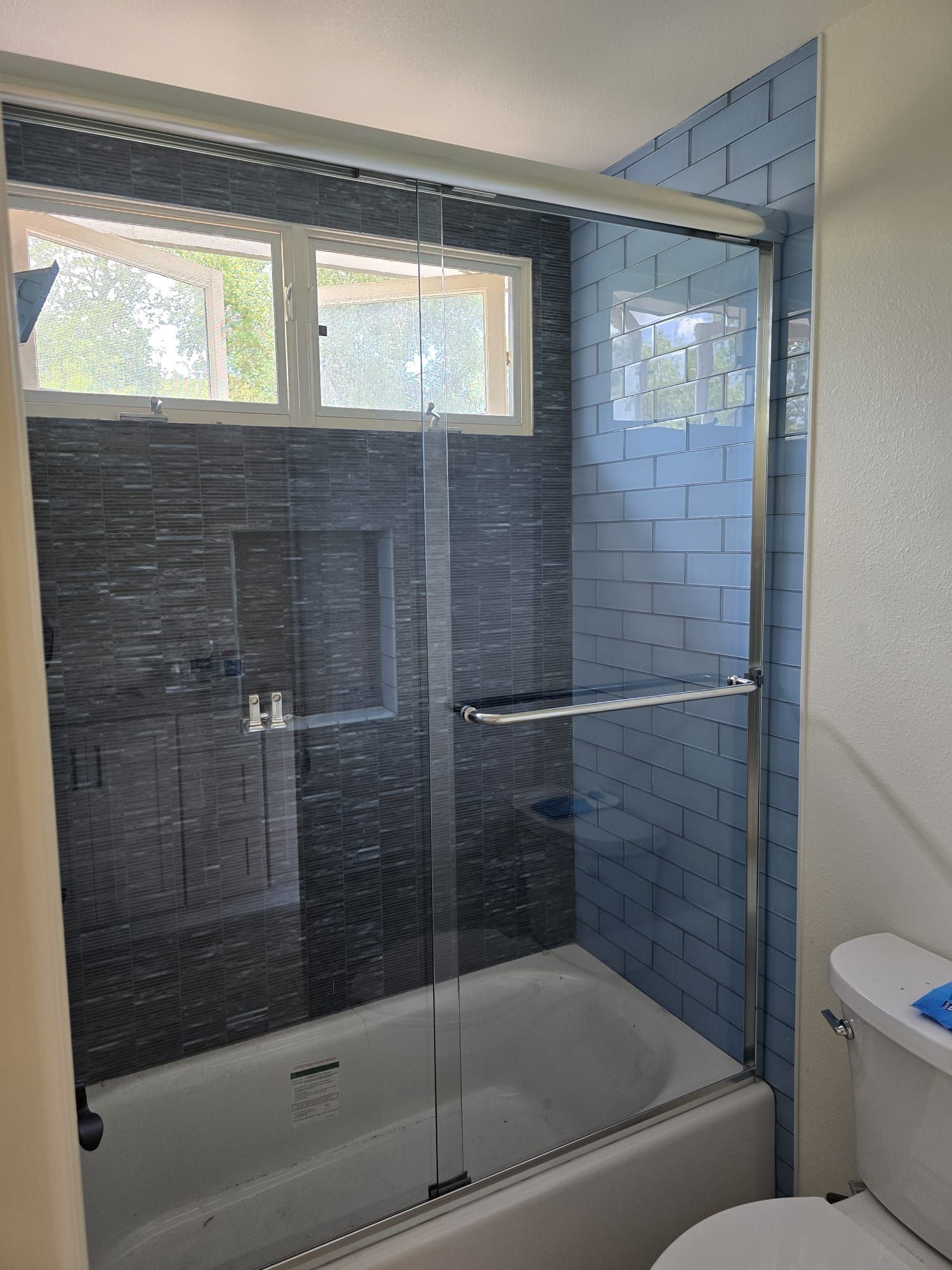 A bathroom featuring a white bathtub with a sliding glass door, dark charcoal tile walls, and a light blue tiled side wall.
