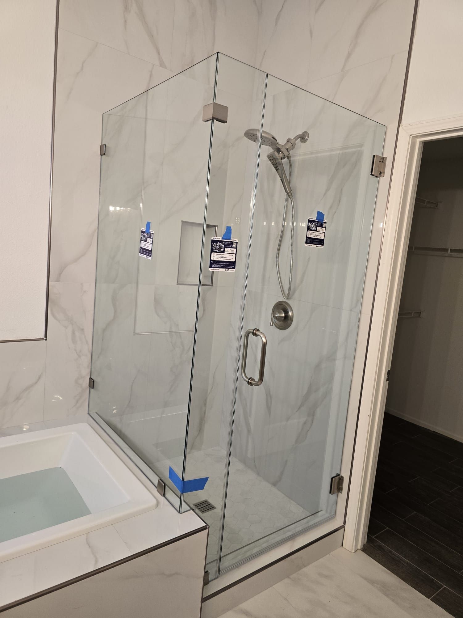 A glass shower enclosure with chrome hardware and a handheld shower head, set against light marble-patterned tile walls.