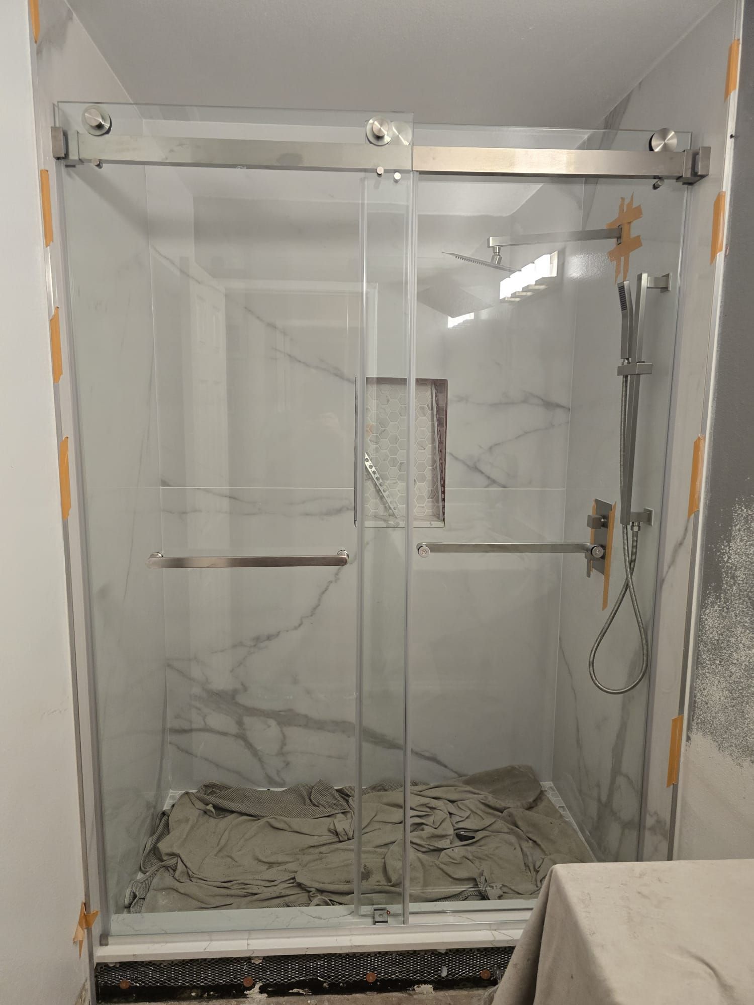 A modern walk-in shower with white marble-patterned tiled walls, a glass sliding door, and a silver handheld showerhead.