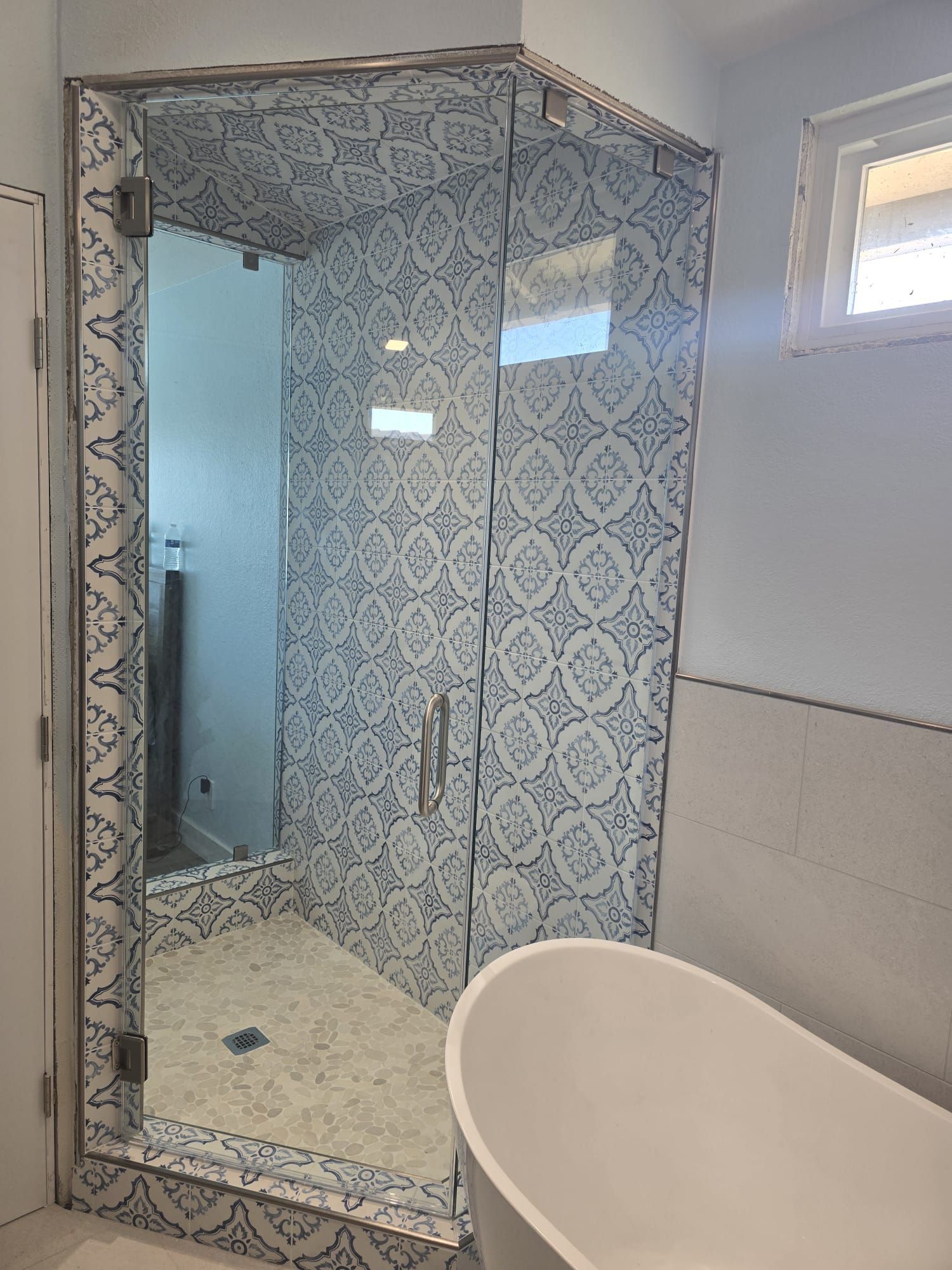 A bathroom shower with patterned blue and white tile walls and a glass door, situated next to a freestanding white bathtub.