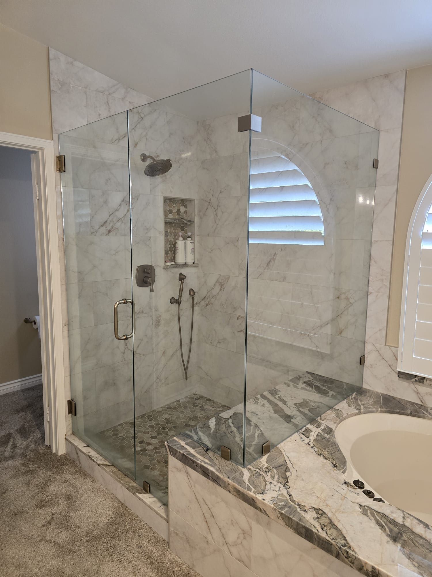 A glass-enclosed shower with marble tile walls and floors, featuring a built-in shower niche and adjacent bathtub.