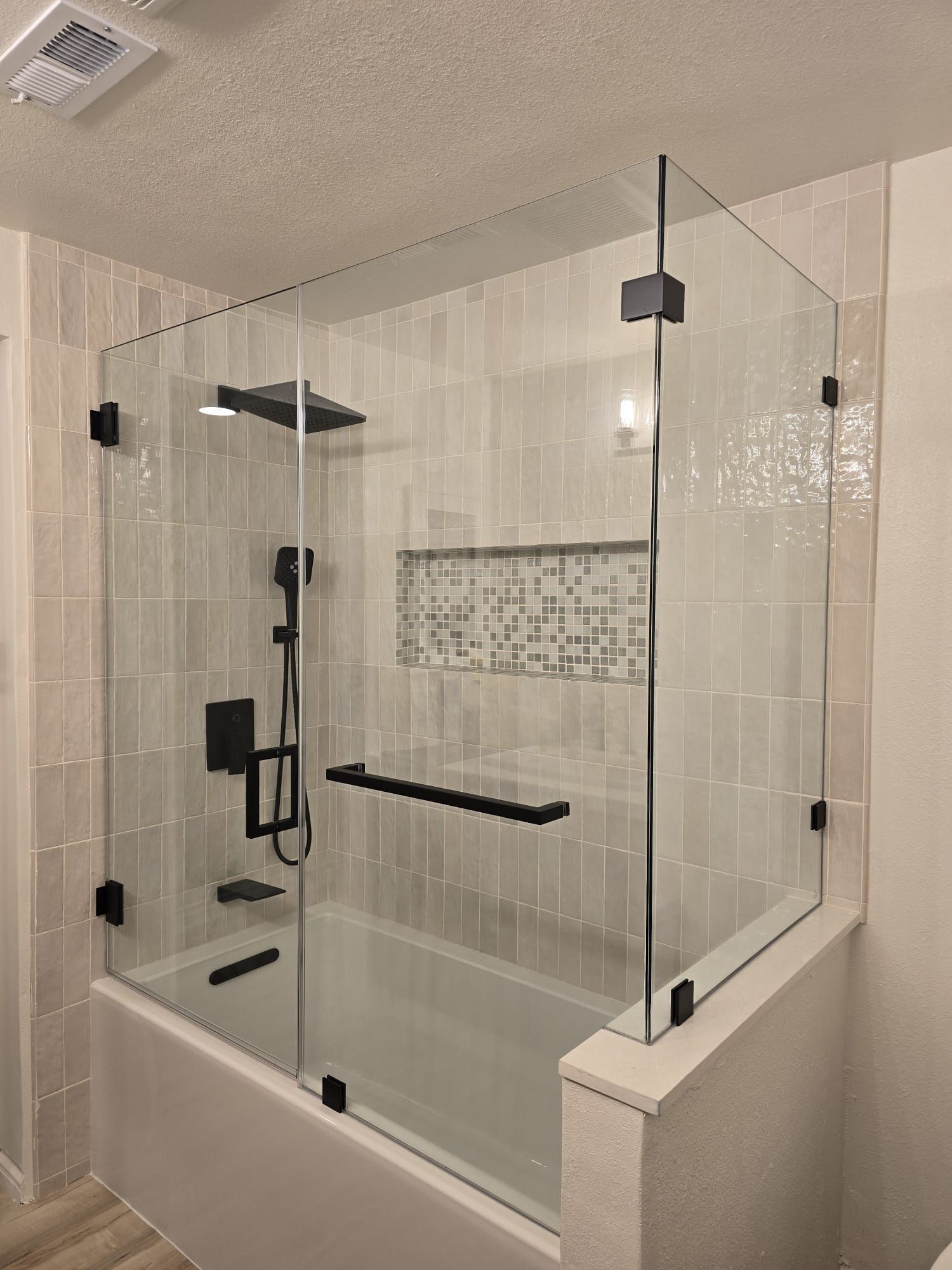A modern bathtub with a clear, frameless glass enclosure and black hardware, set against light-colored tiled walls.