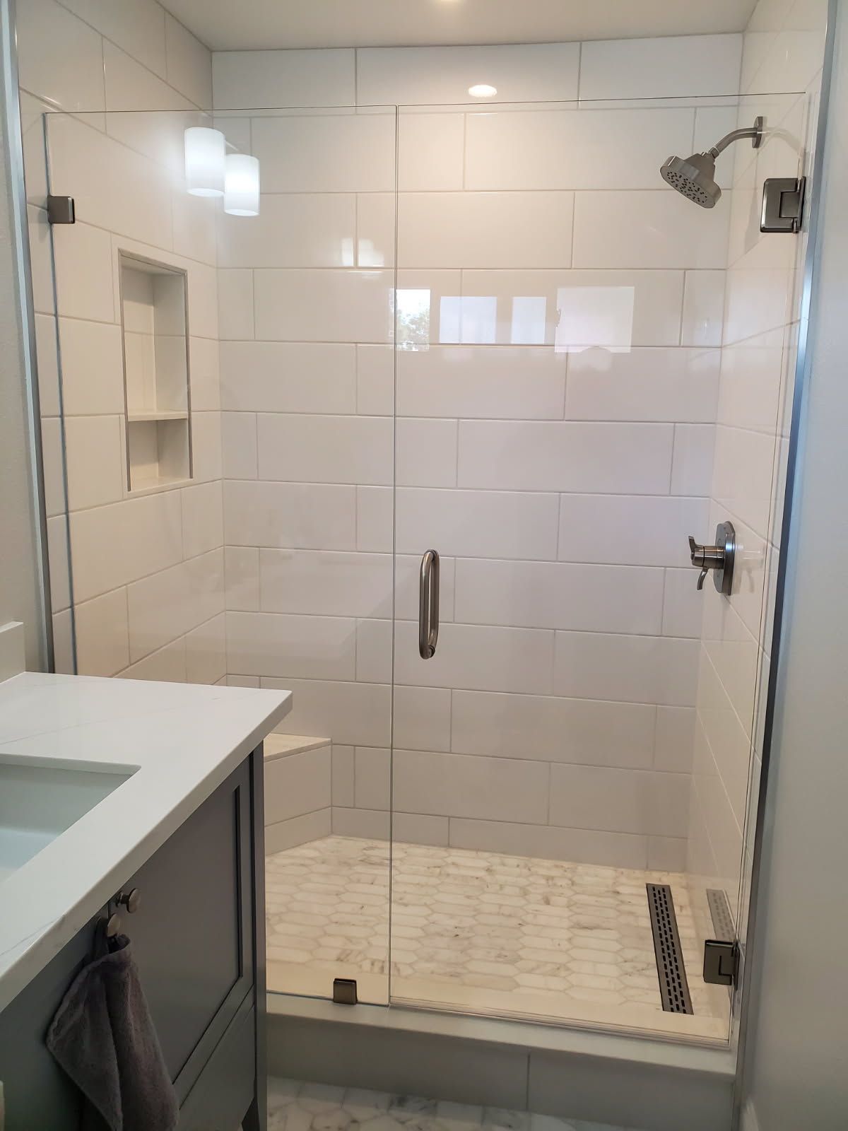 A modern walk-in shower with white subway tiles, a glass door, a built-in shelf, stone flooring, and a showerhead.