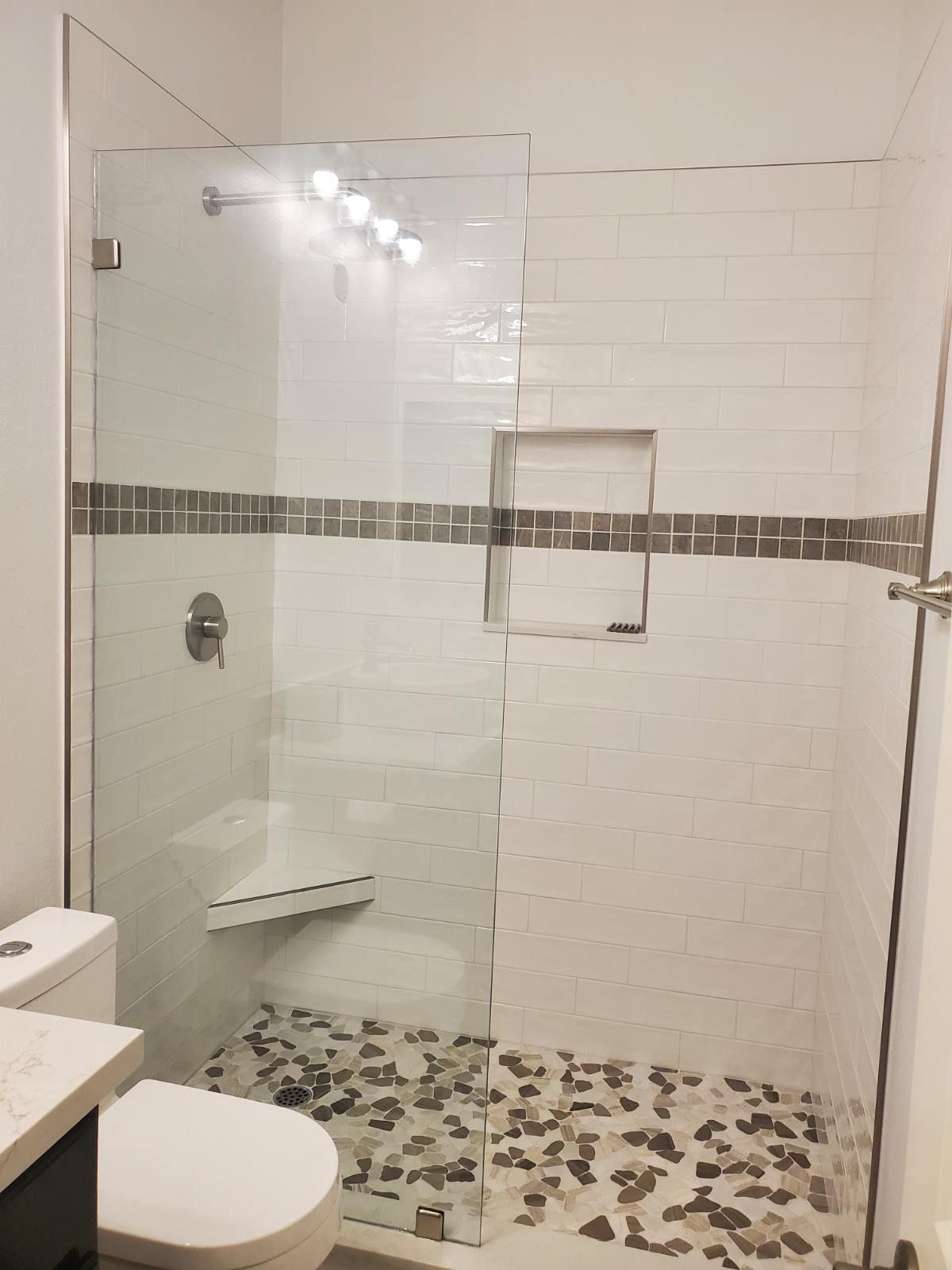A modern walk-in shower with white subway tiles, a mosaic accent strip, a stone pebble floor, and a glass partition.