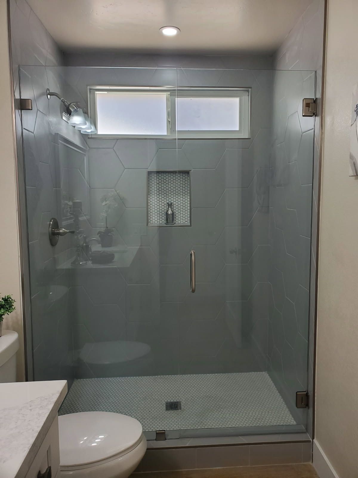 A modern walk-in shower with gray hexagonal wall tiles, a small recessed shelf, and a glass door, adjacent to a toilet.