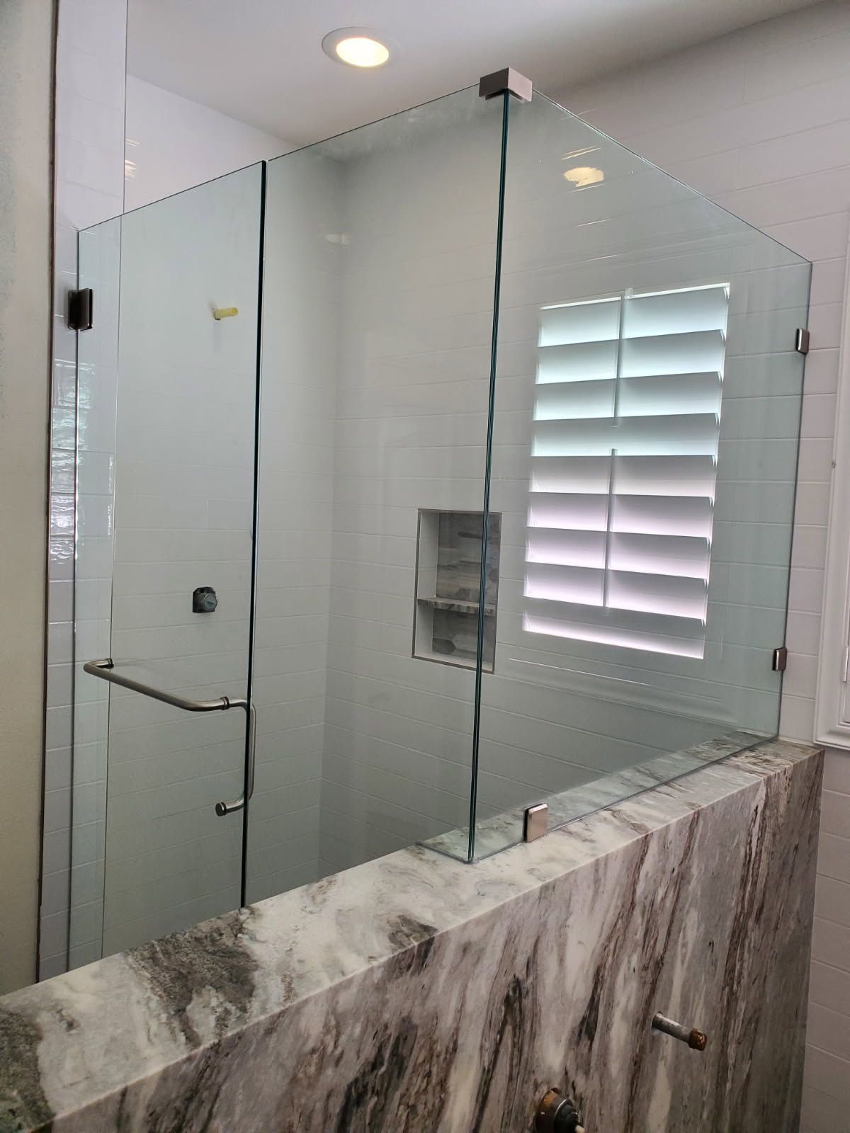 A frameless glass shower enclosure sits atop a marble-topped half wall in a bathroom with white shutters.