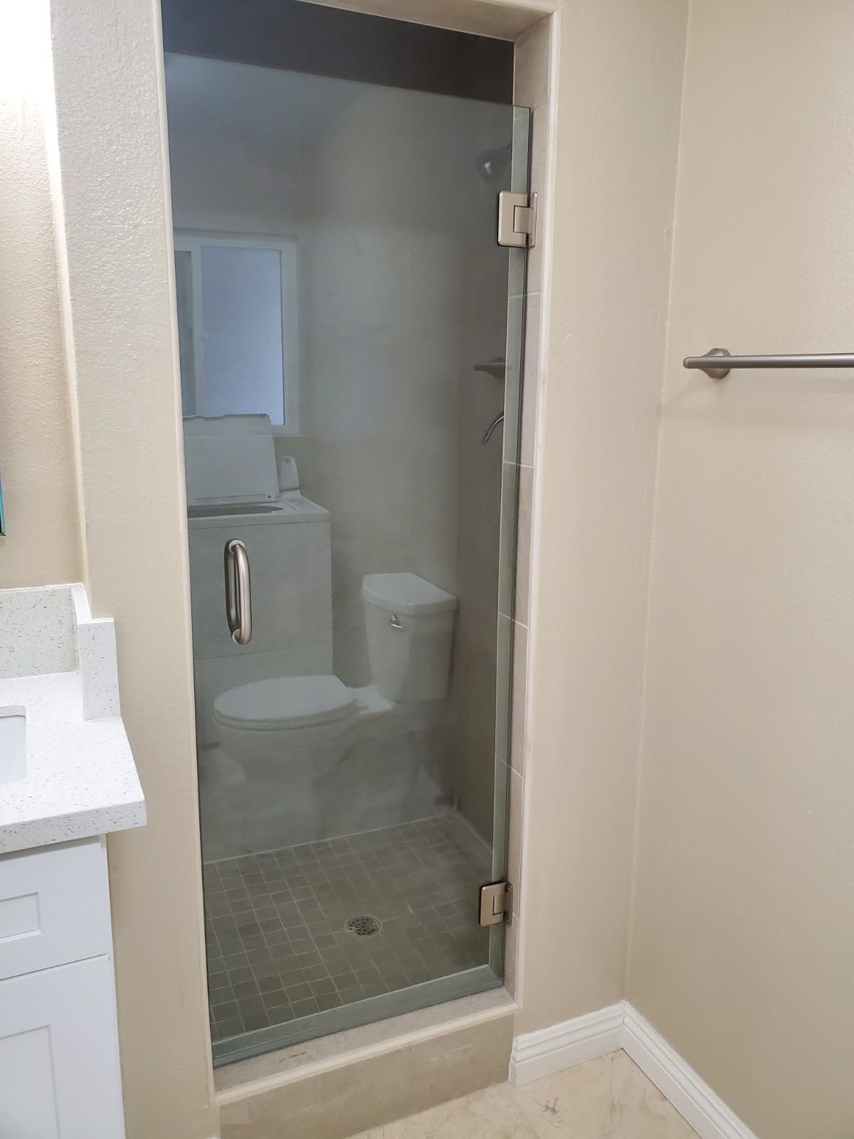 A bathroom glass shower door with a metal handle and hinges, revealing a toilet inside the tiled shower stall.