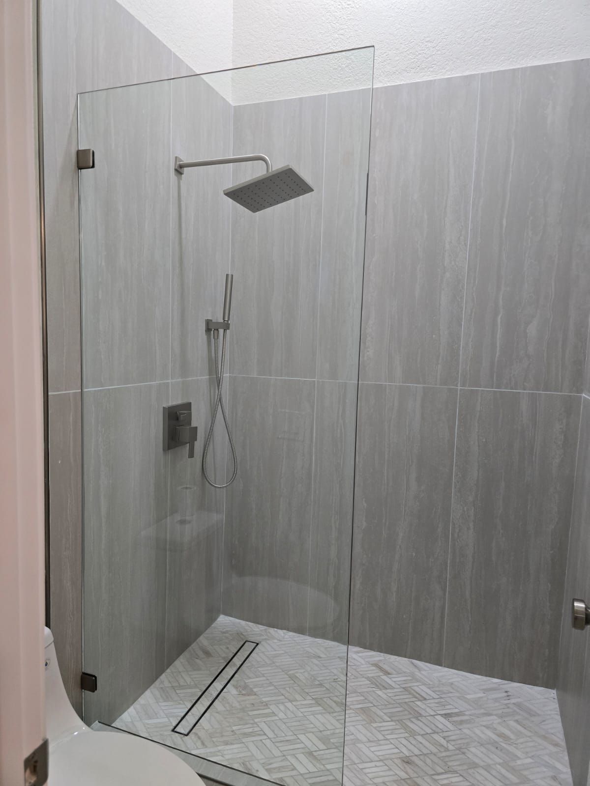 Modern walk-in shower with gray tiled walls, a glass partition, rainfall showerhead, and white marble-patterned floor.
