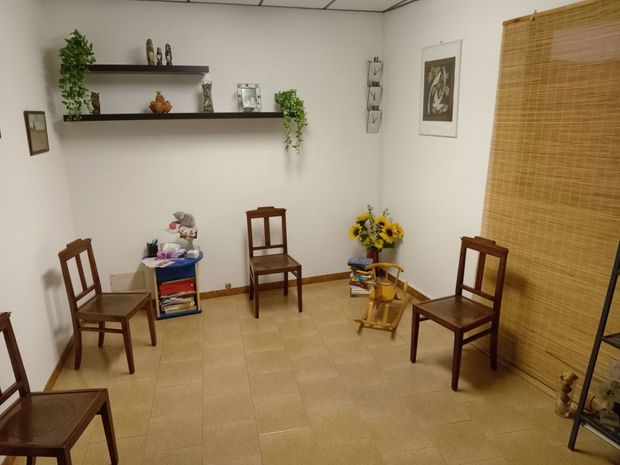 Piccola sala d'attesa con sedie in legno, scaffali, quadri incorniciati e una tenda a rullo in tessuto.
