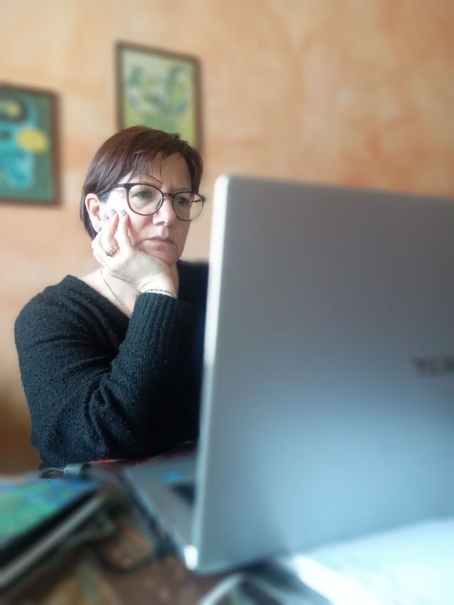 Una persona è seduta al computer portatile in una stanza accogliente, con il mento appoggiato alla mano, e alle pareti sono appesi quadri incorniciati.