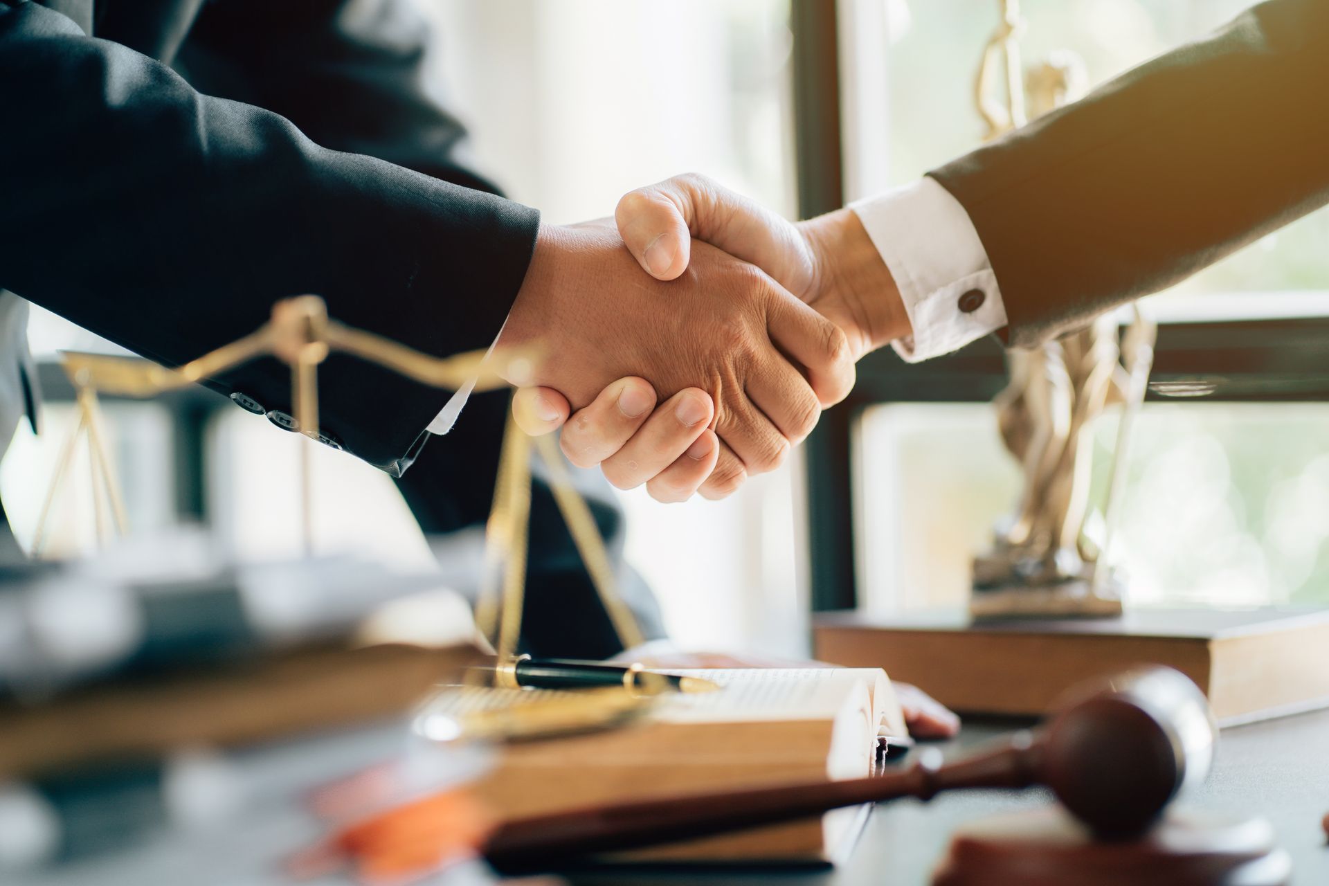 Two people shaking hands over a desk with legal items, including a gavel, scales of justice, and law