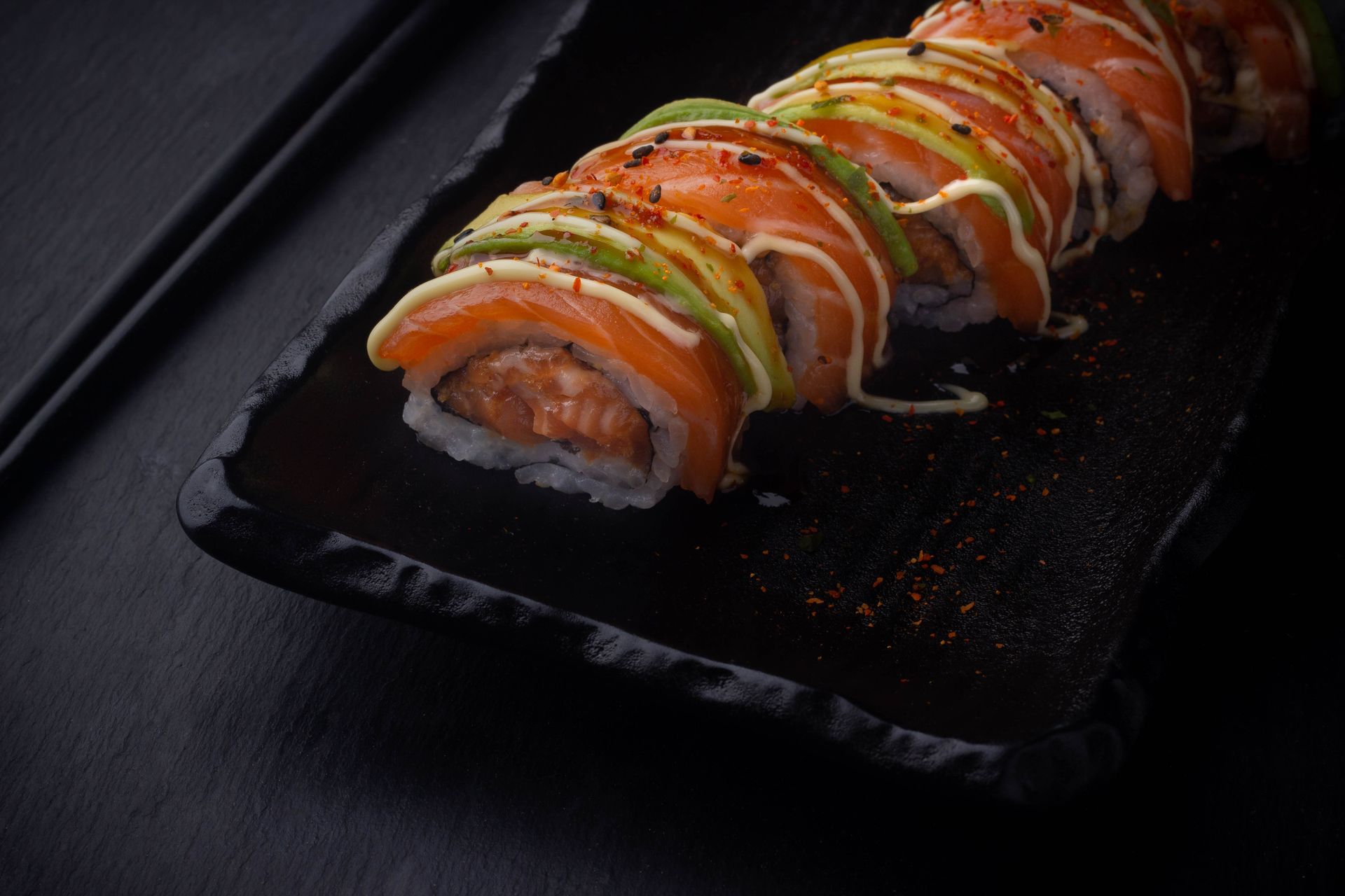 Sushi Restaurant | Finding Sushi | Sushi rolls topped with avocado, salmon, and sauce on a black rectangular plate