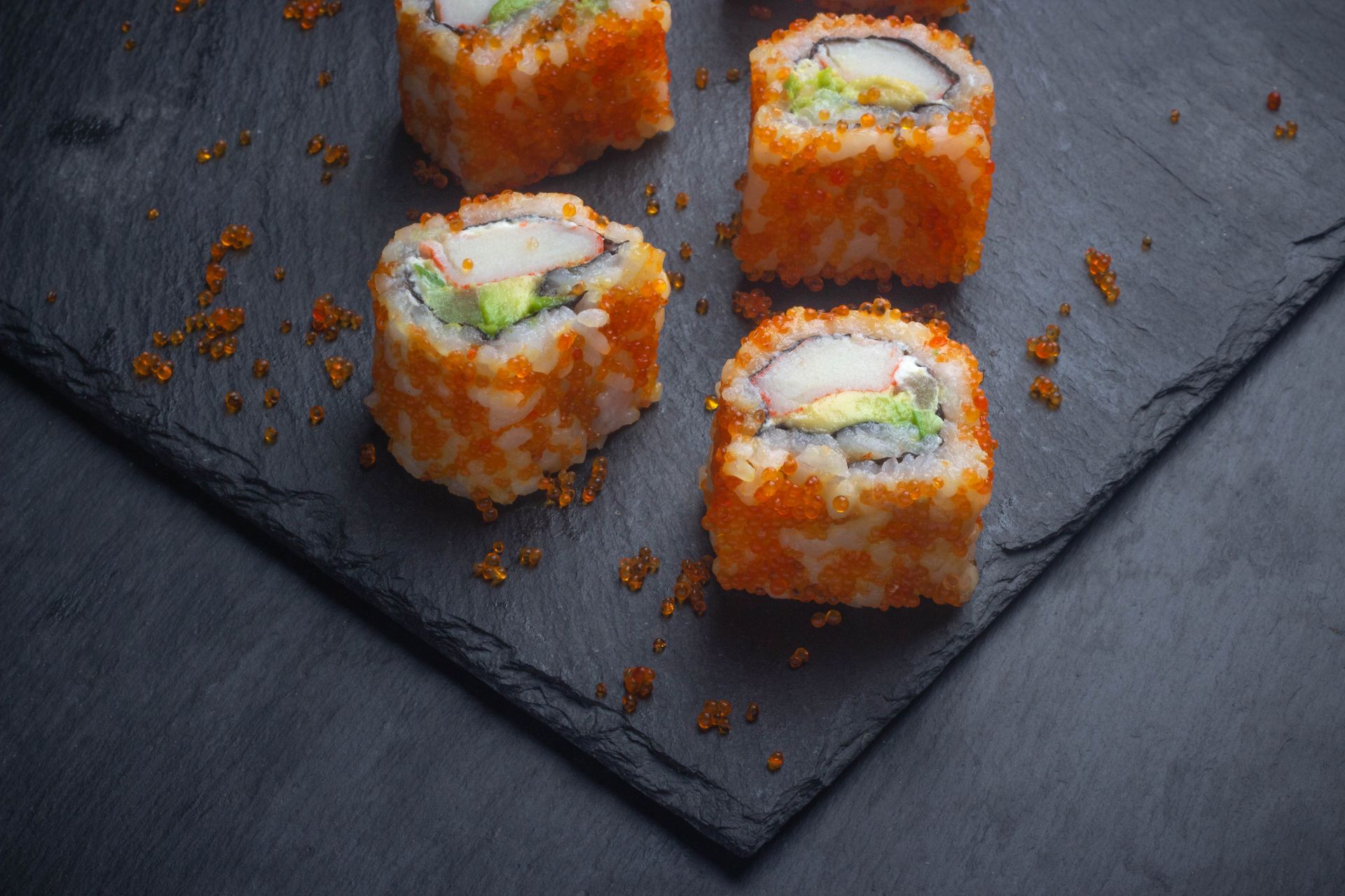 Sushi Restaurant | Finding Sushi | Sushi rolls on a dark slate plate, topped with orange roe and garnished with green herbs.