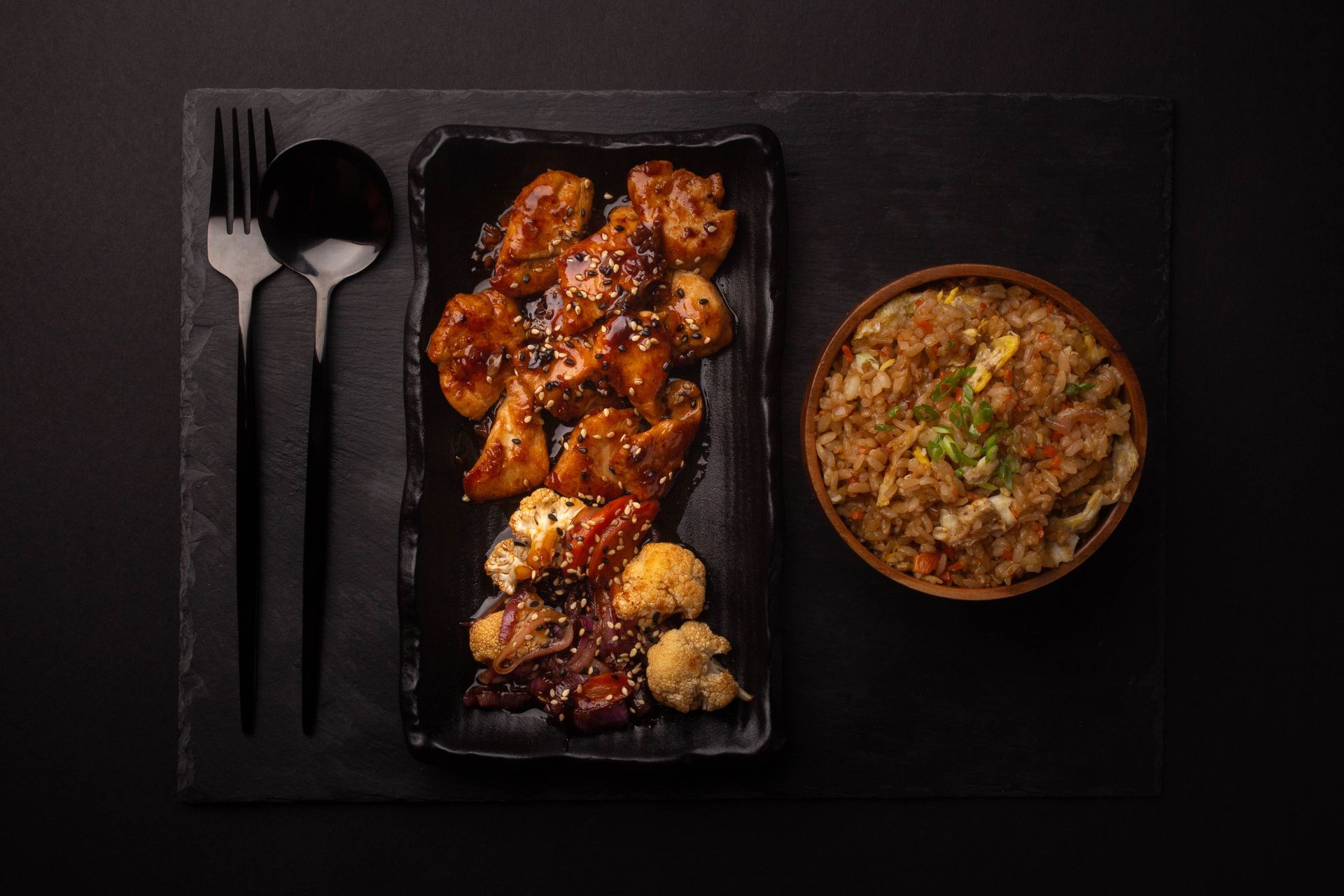 Black tray with glazed chicken skewers and rice, beside a fork, spoon, and knife on a dark table