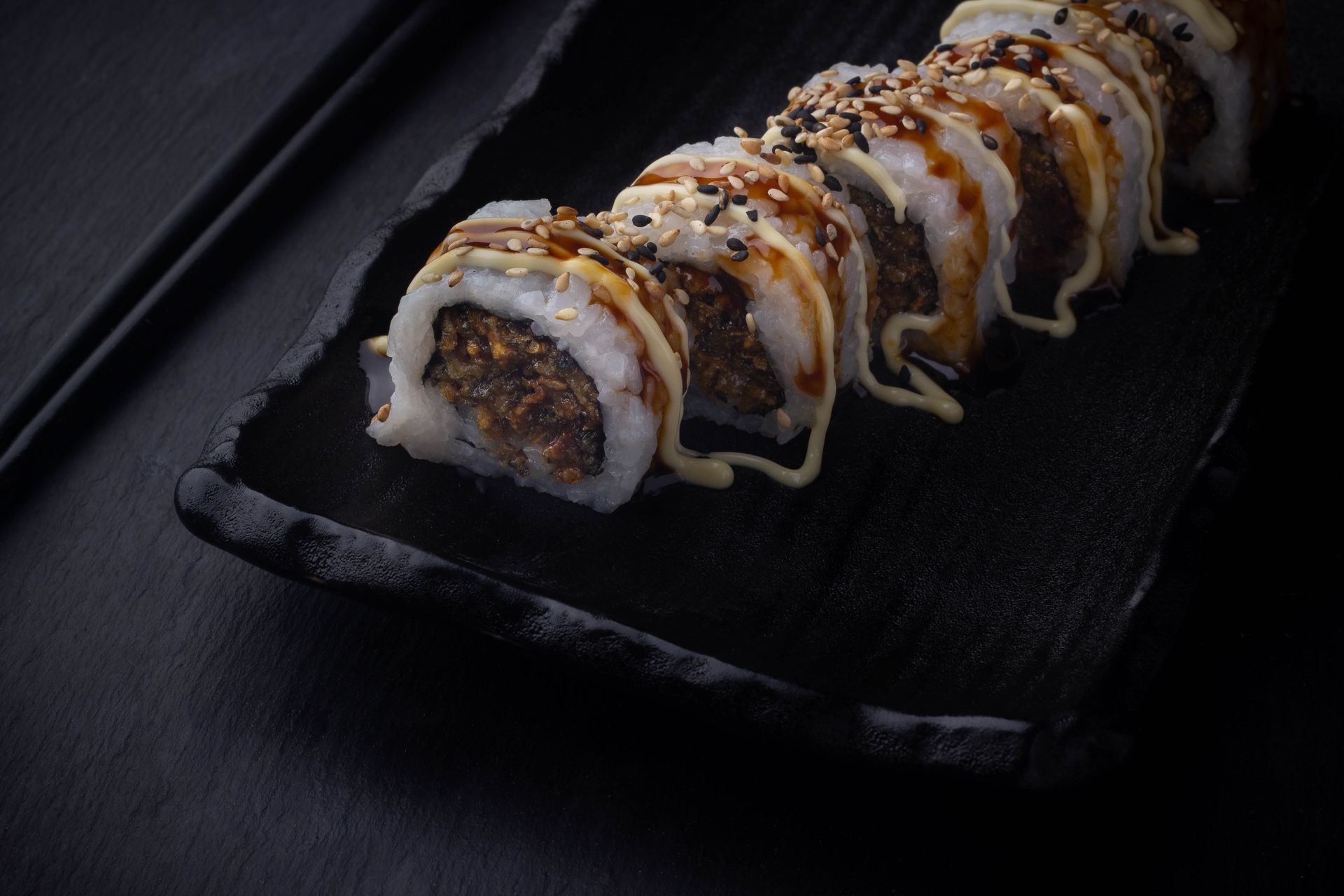 Sushi Restaurant | Finding Sushi | Sushi roll topped with sauce and sesame seeds on a black serving plate