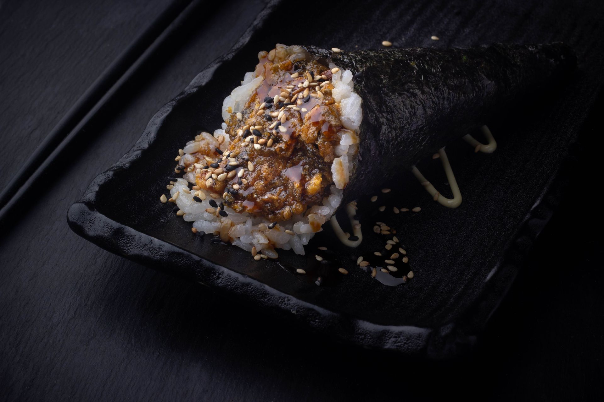 Sushi Restaurant | Finding Sushi | Sushi roll topped with sesame seeds on a black plate, with drizzle on the side