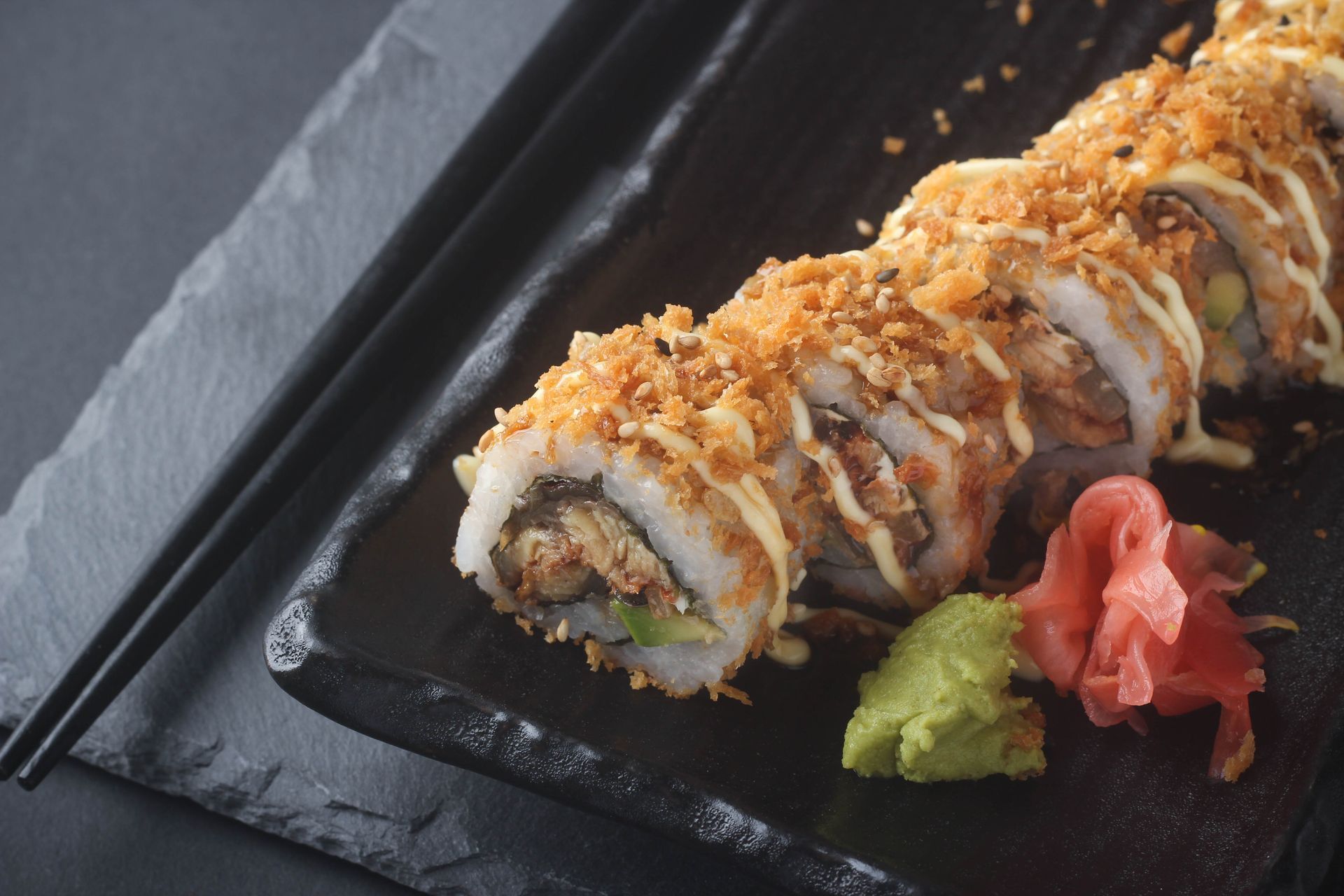 Sushi Restaurant | Finding Sushi | Sushi roll topped with sauce and crumbs on a black plate, with chopsticks, wasabi,