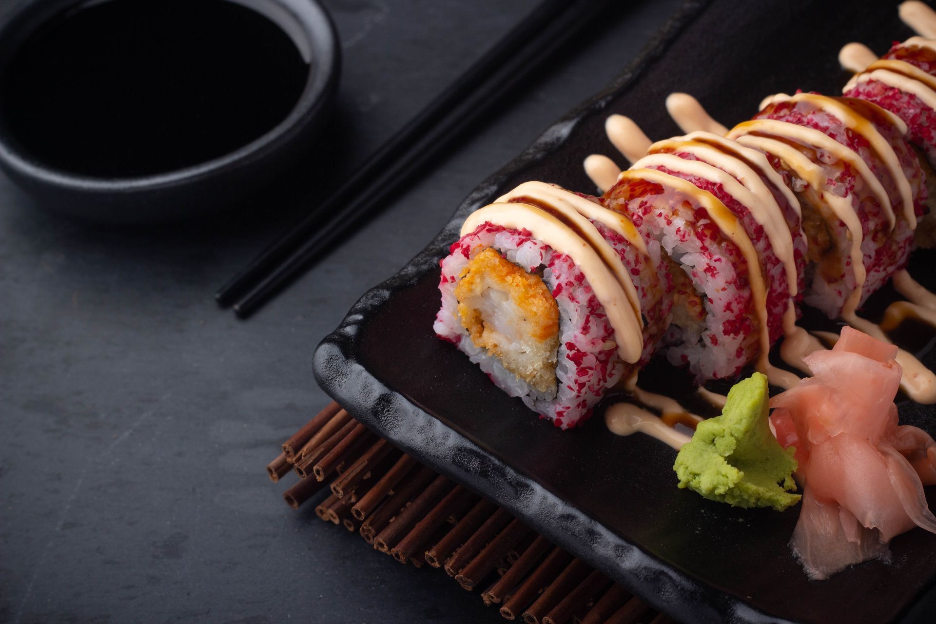 Sushi rolls on a black plate with wasabi, pickled ginger, chopsticks, and soy sauce