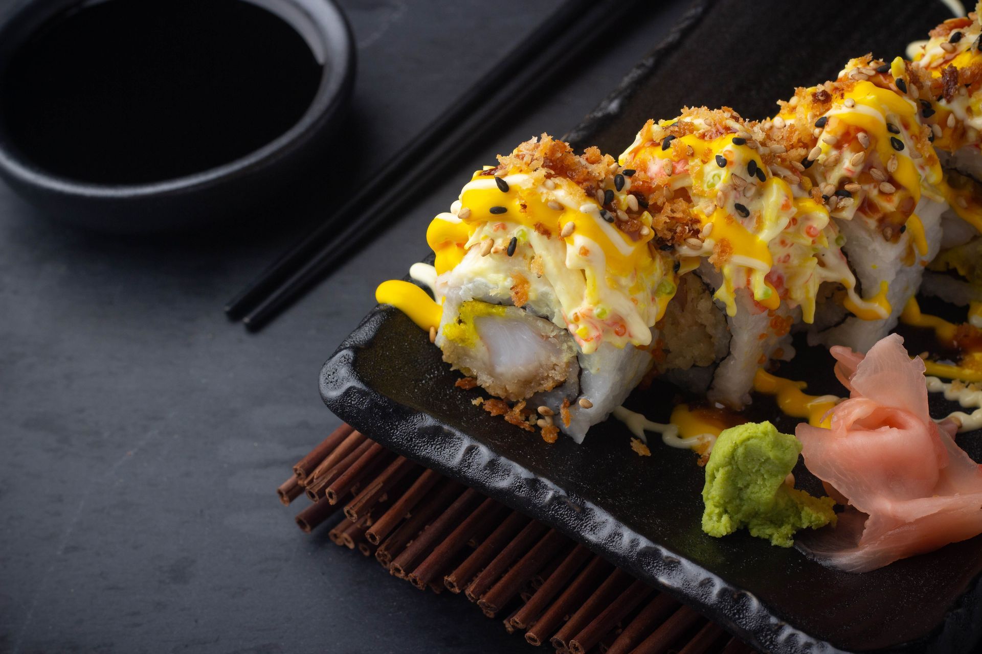 Sushi Restaurant | Finding Sushi | Sushi rolls topped with sauce on a black plate, with chopsticks, soy sauce, wasabi