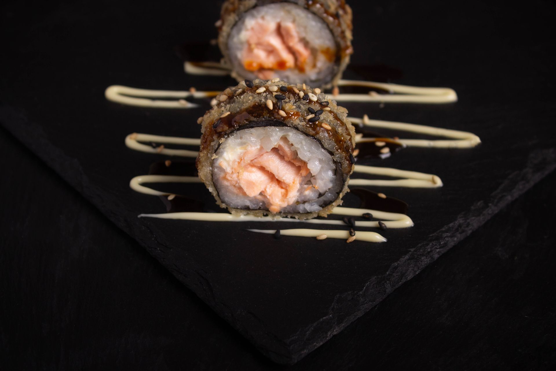 Sushi Restaurant | Finding Sushi | Sushi rolls on a black slate plate, drizzled with sauce and served with salmon topping