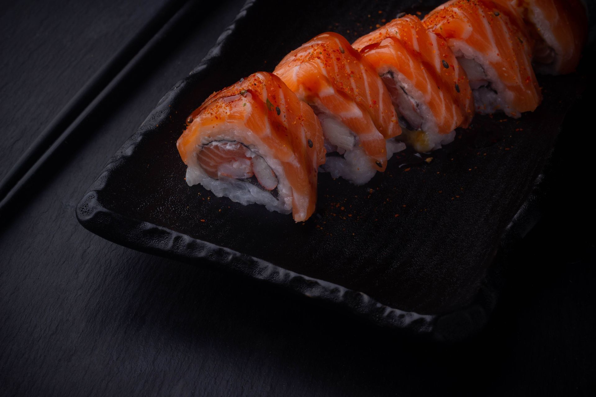 Sushi Restaurant | Finding Sushi | Sushi rolls topped with orange fish on a black plate against a dark background