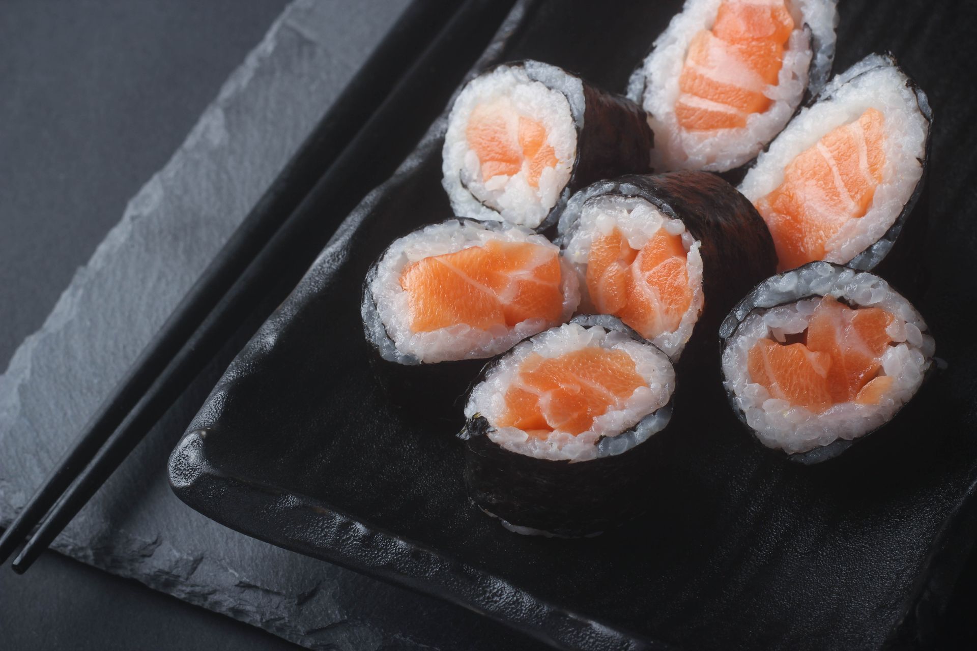 Sushi Restaurant | Finding Sushi | Salmon sushi rolls on a black plate with chopsticks on a dark surface
