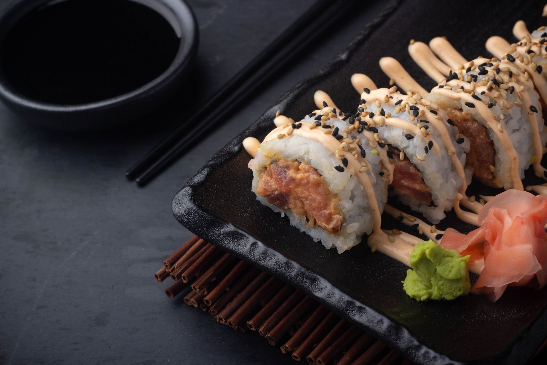 Sushi Restaurant | Finding Sushi | Sushi rolls drizzled with sauce on a black plate, with soy sauce, chopsticks, ginger