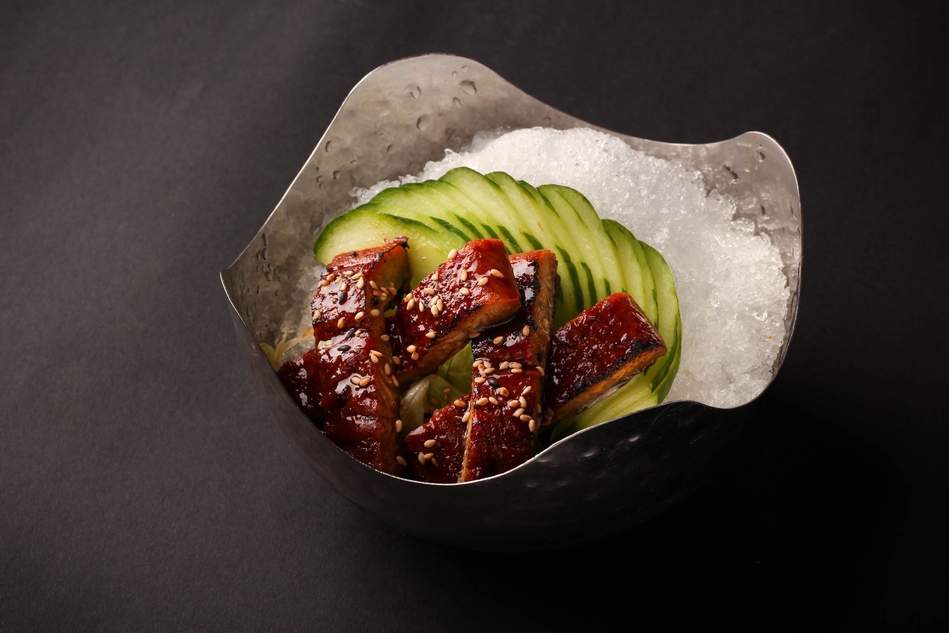 Glazed meat slices with sesame seeds arranged over cucumber slices in a dark bowl