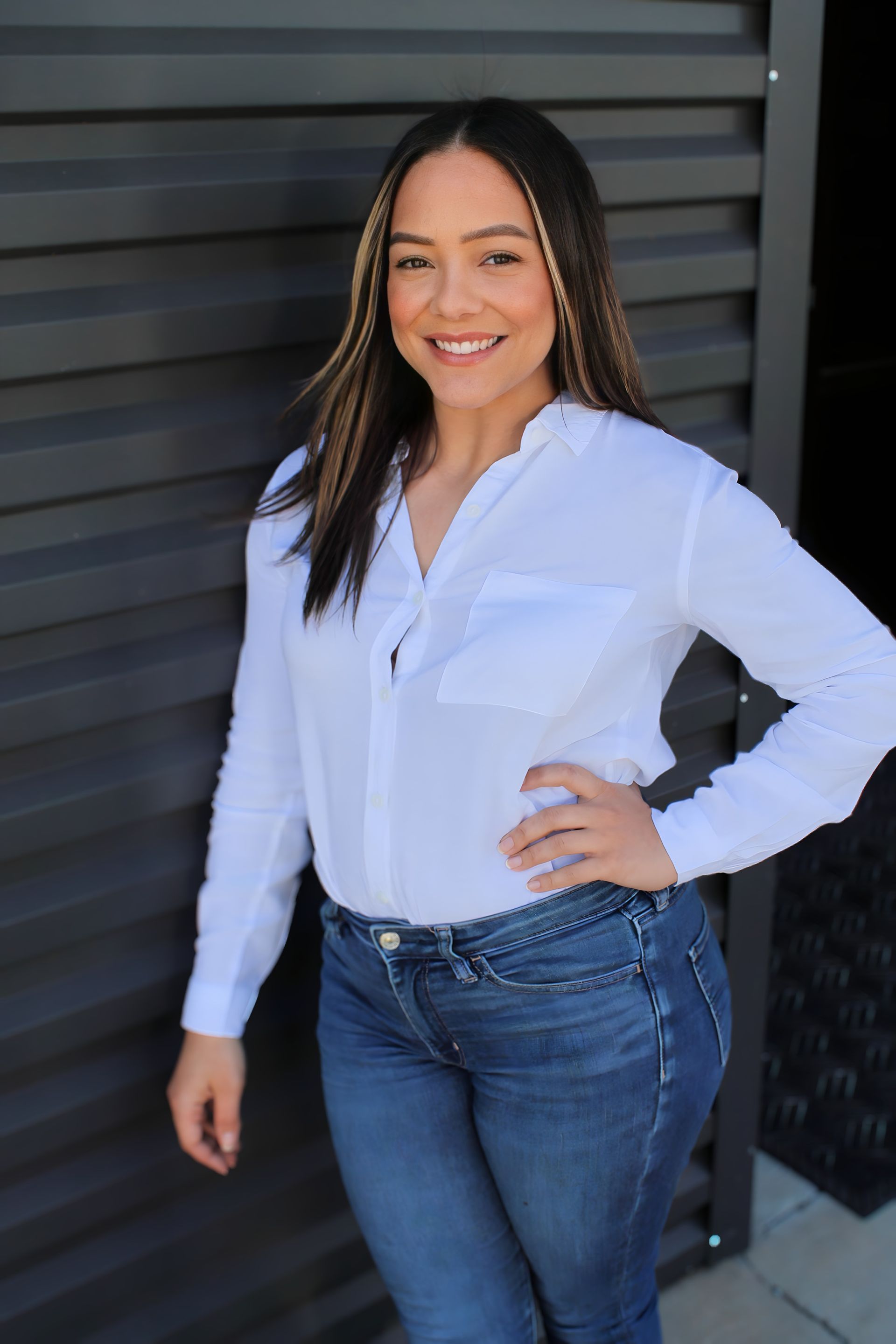 A woman wearing a white shirt and blue jeans is standing in front of a wall.