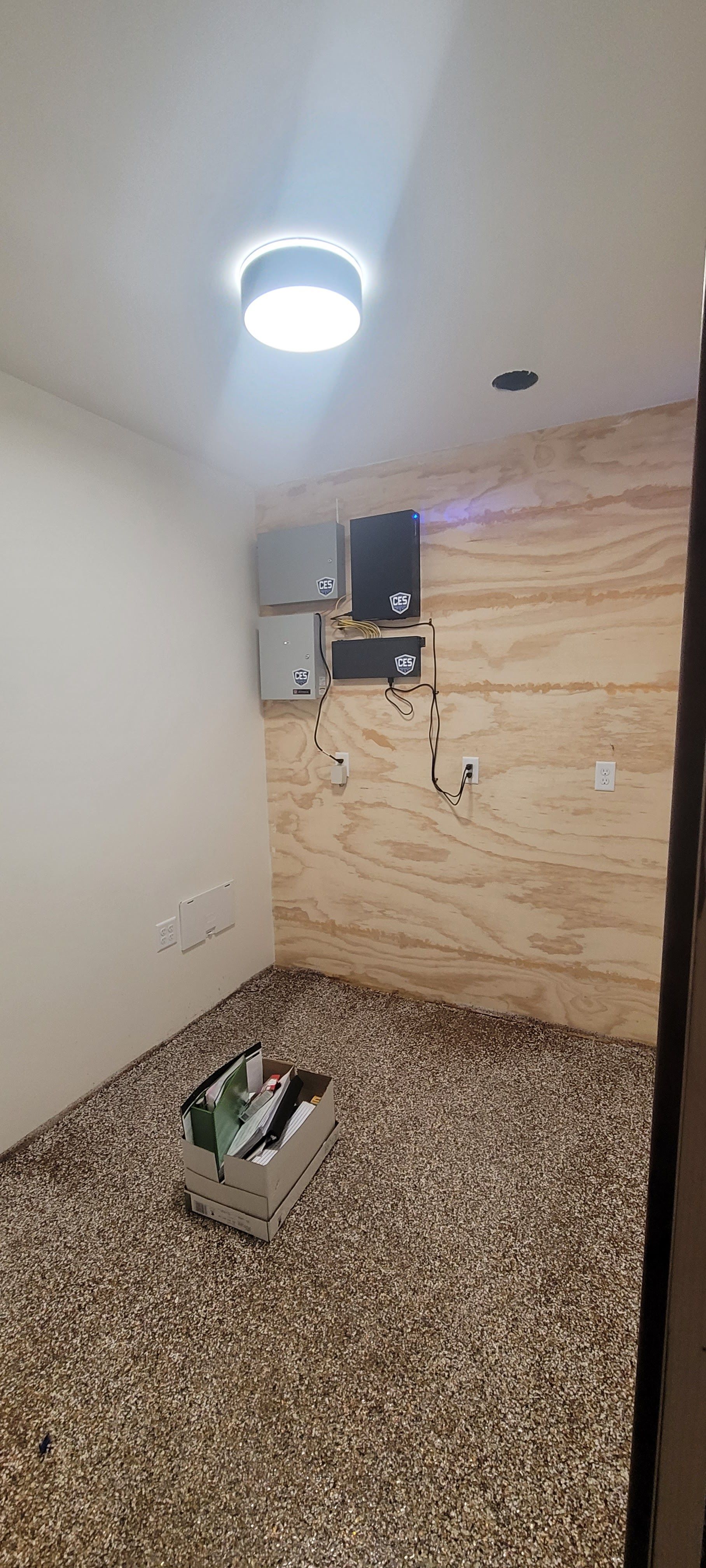 Small room under renovation with plywood wall, ceiling light, wall-mounted TV, and tools on a speckled floor.