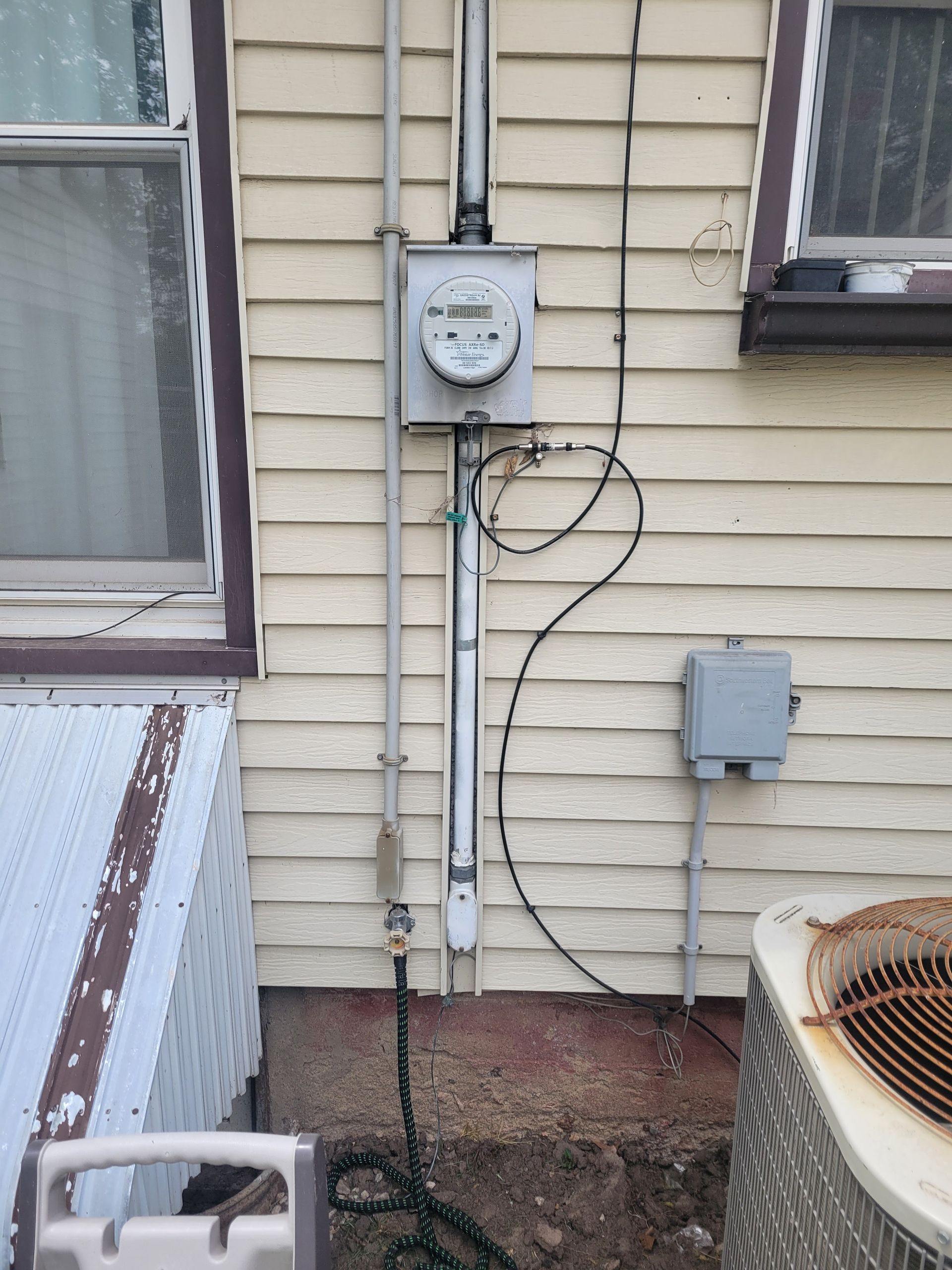Exterior house wall with utility meter, cables, and electrical boxes beside windows and a window AC unit
