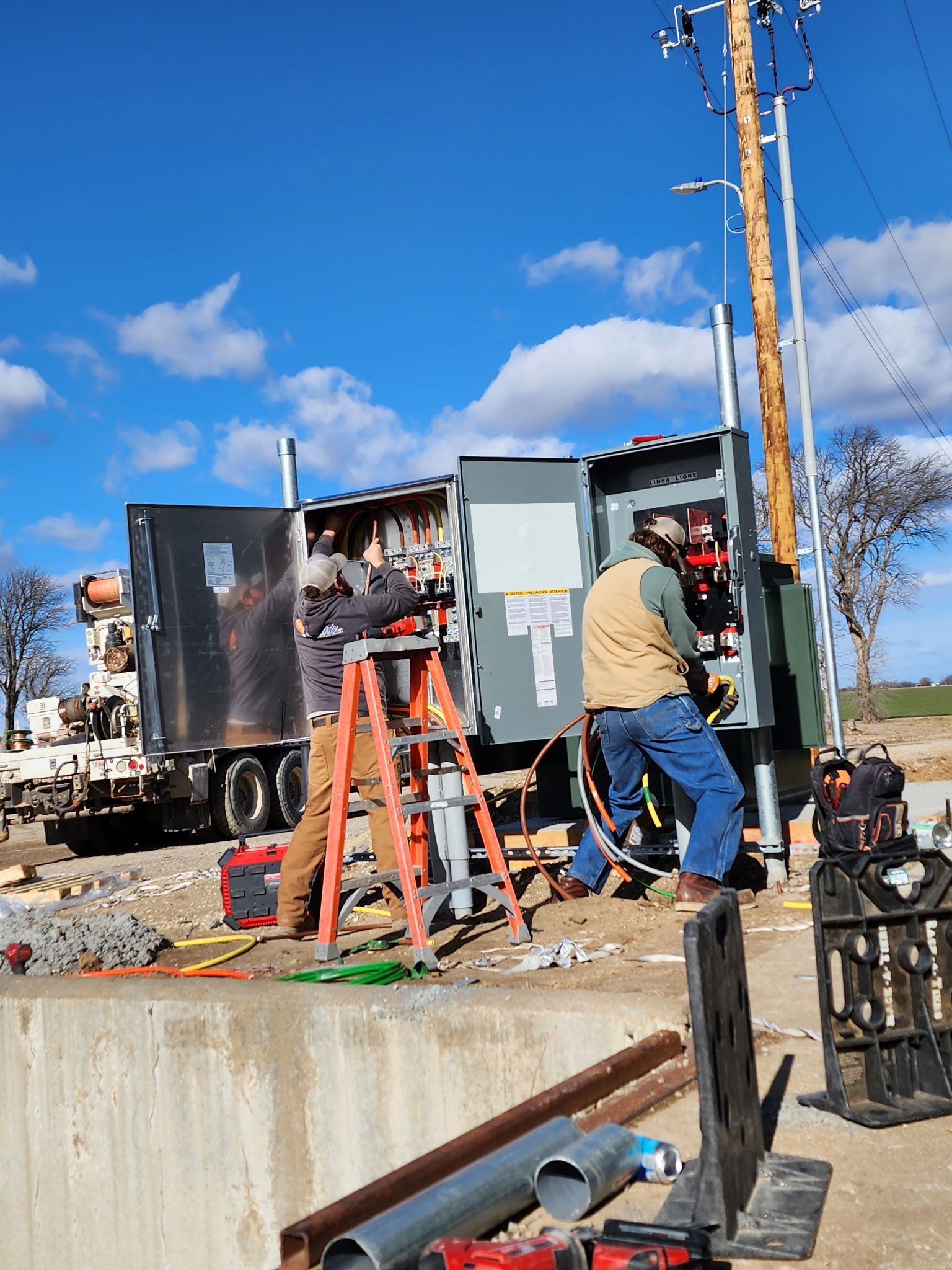 Construction workers at a roadside utility site with equipment and a ladder under a blue sky.