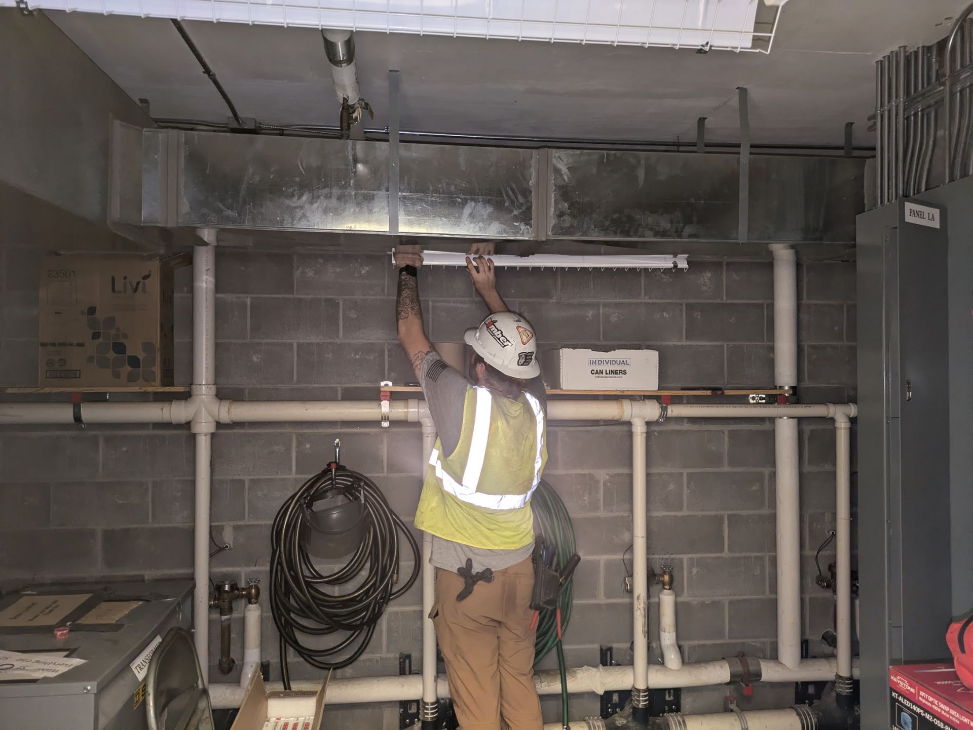 Worker in a safety vest adjusts ceiling pipes in an industrial utility room.