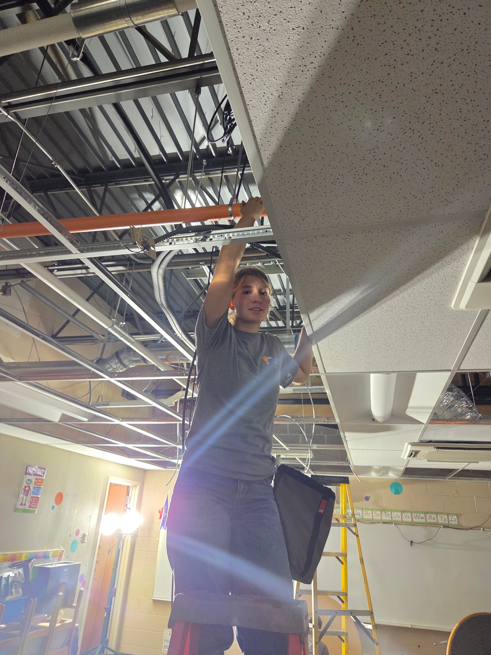 Worker on a ladder adjusting ceiling fixtures in a room under construction.