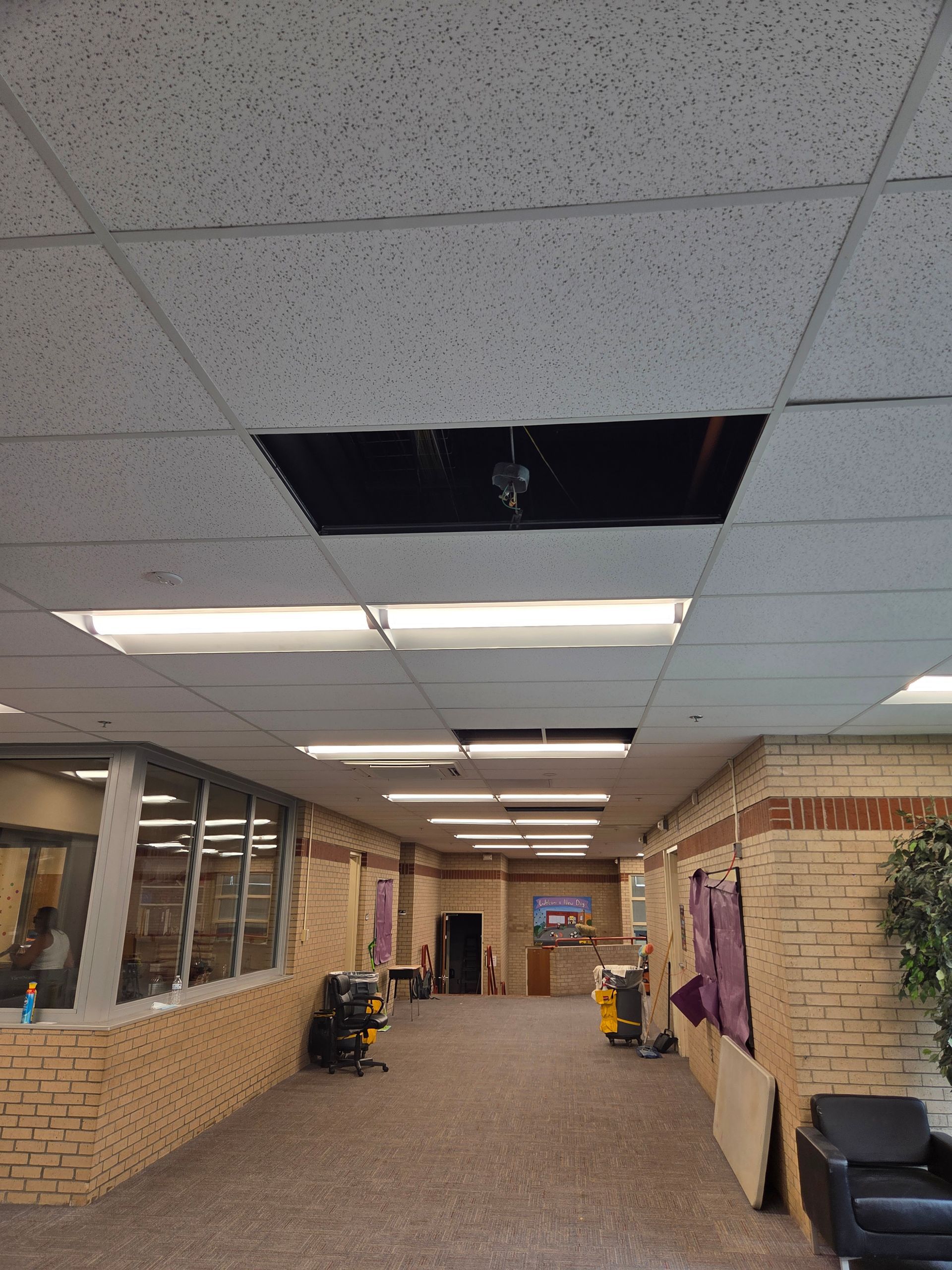 Long hallway with drop ceiling, a damaged ceiling tile, brick walls, windows, and chairs along the right side