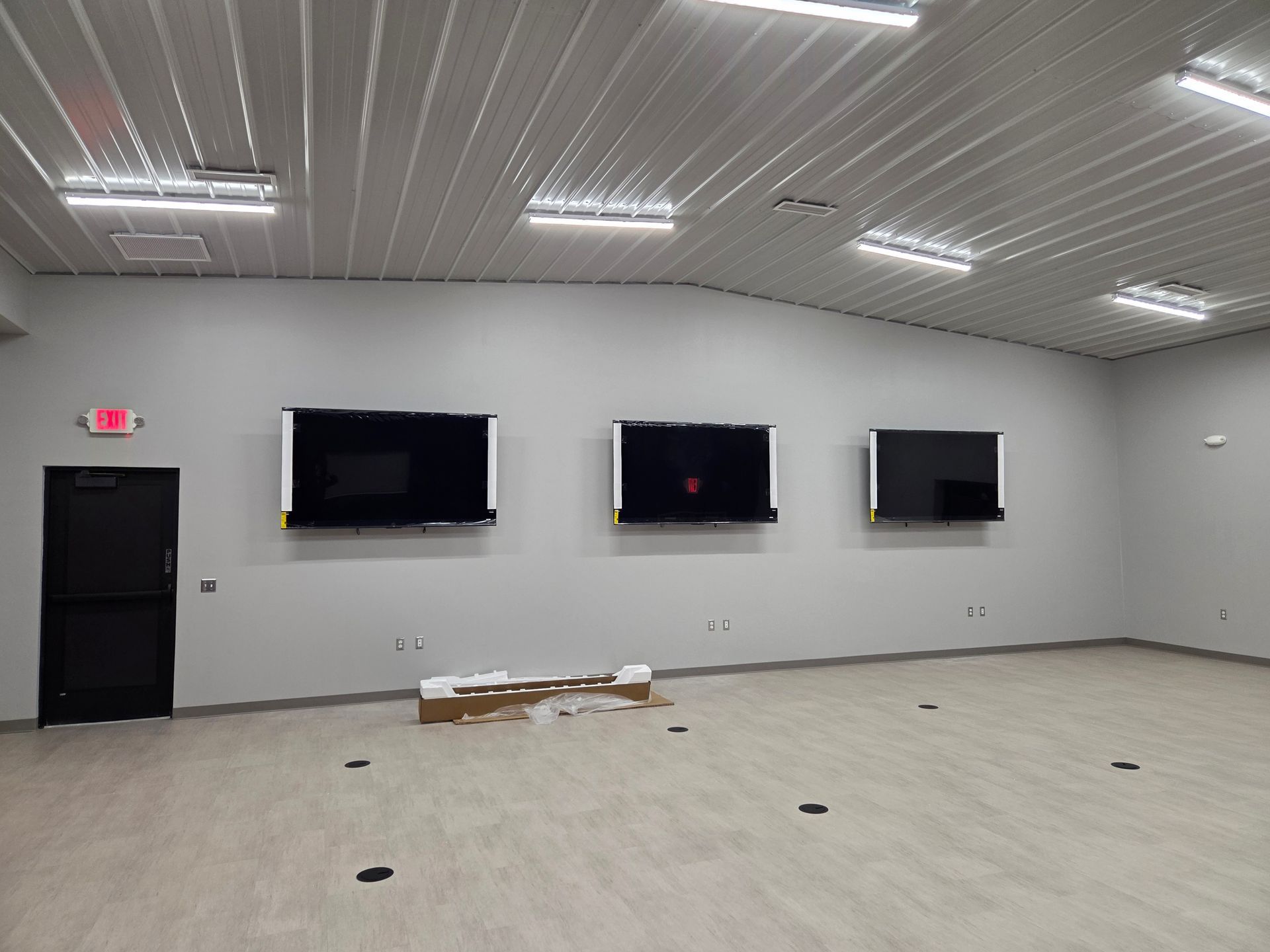 Empty room with three wall-mounted TVs, white ceiling lights, and a black door
